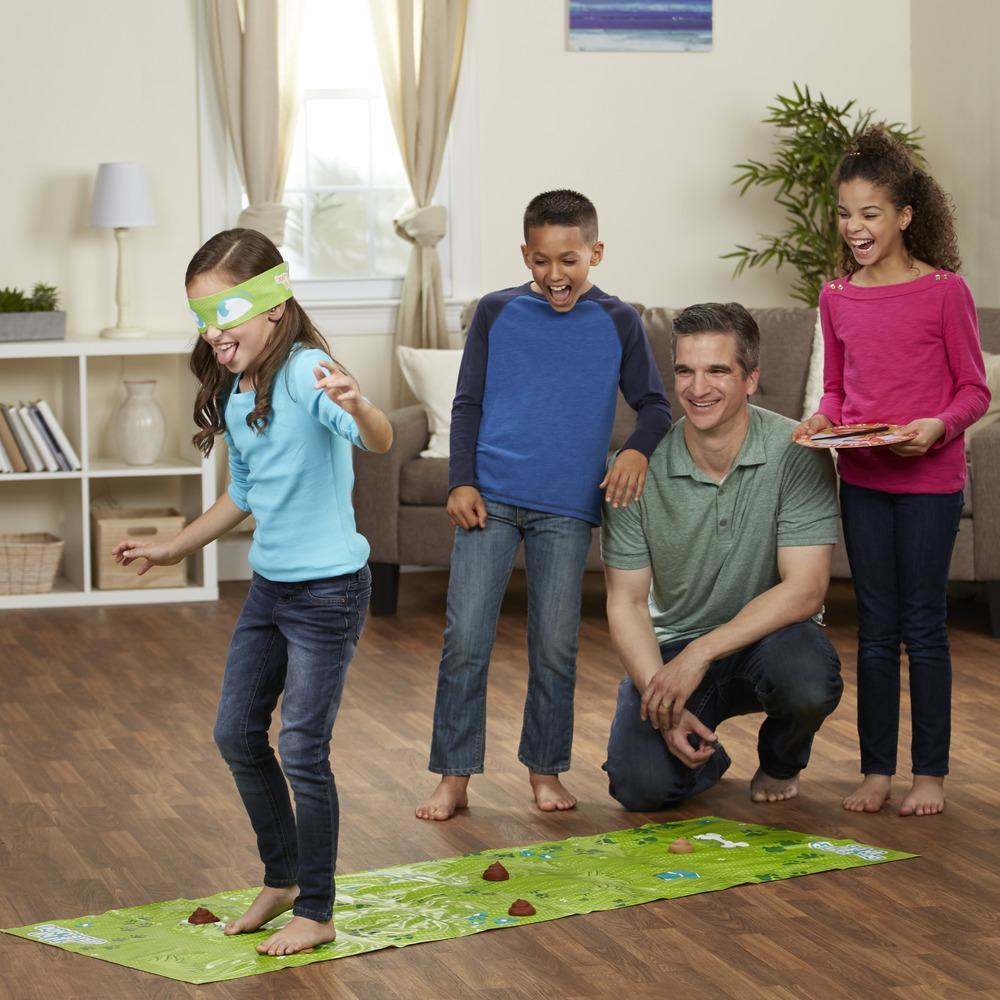 Dzieci grają w grę. Jedno ma zawiązane oczy, inni się uśmiechają, stoją na zielonej macie z brązowymi plamami.