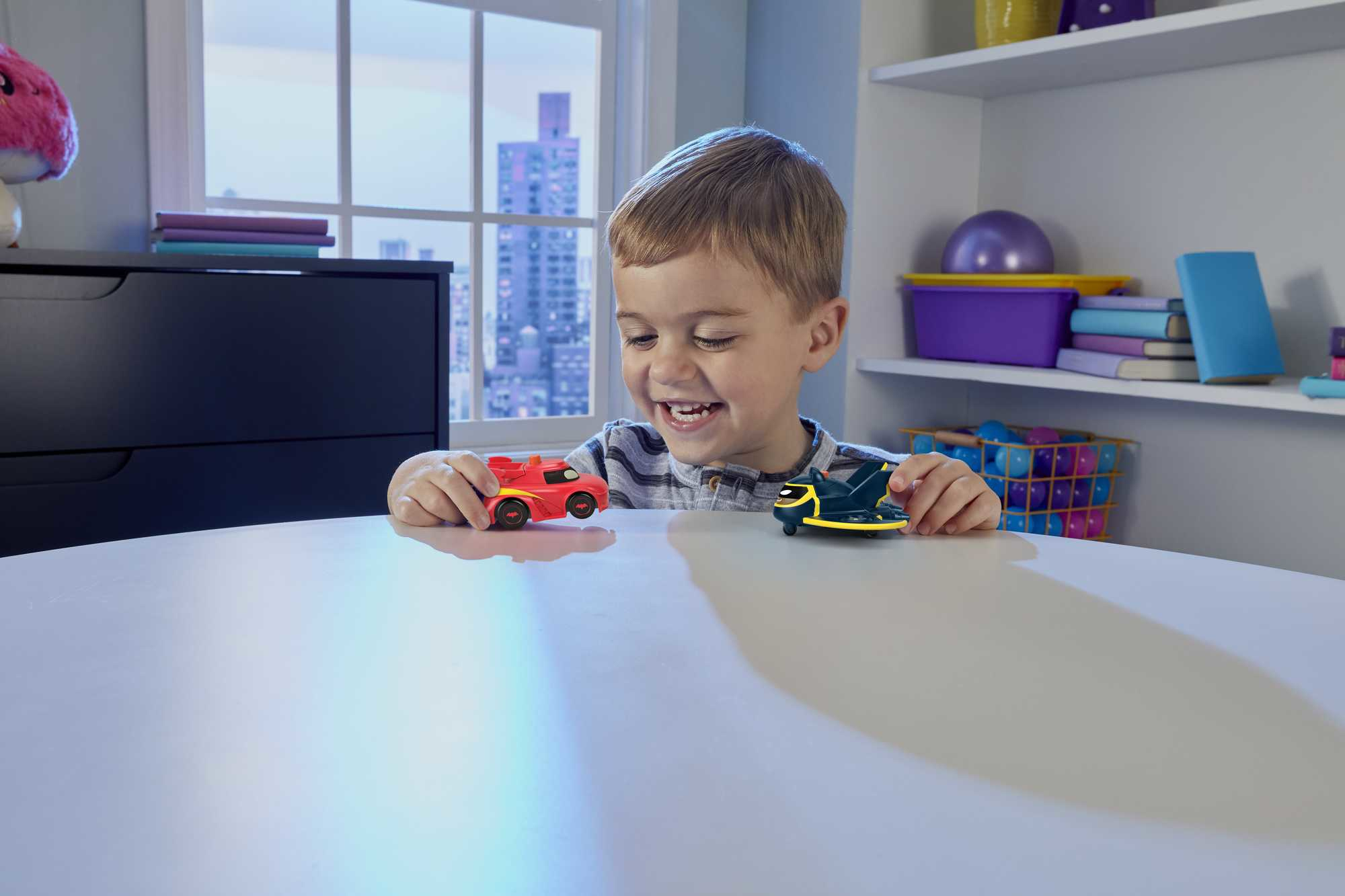 Mały chłopiec uśmiecha się, bawiąc się dwoma samochodzikami na białym stole.