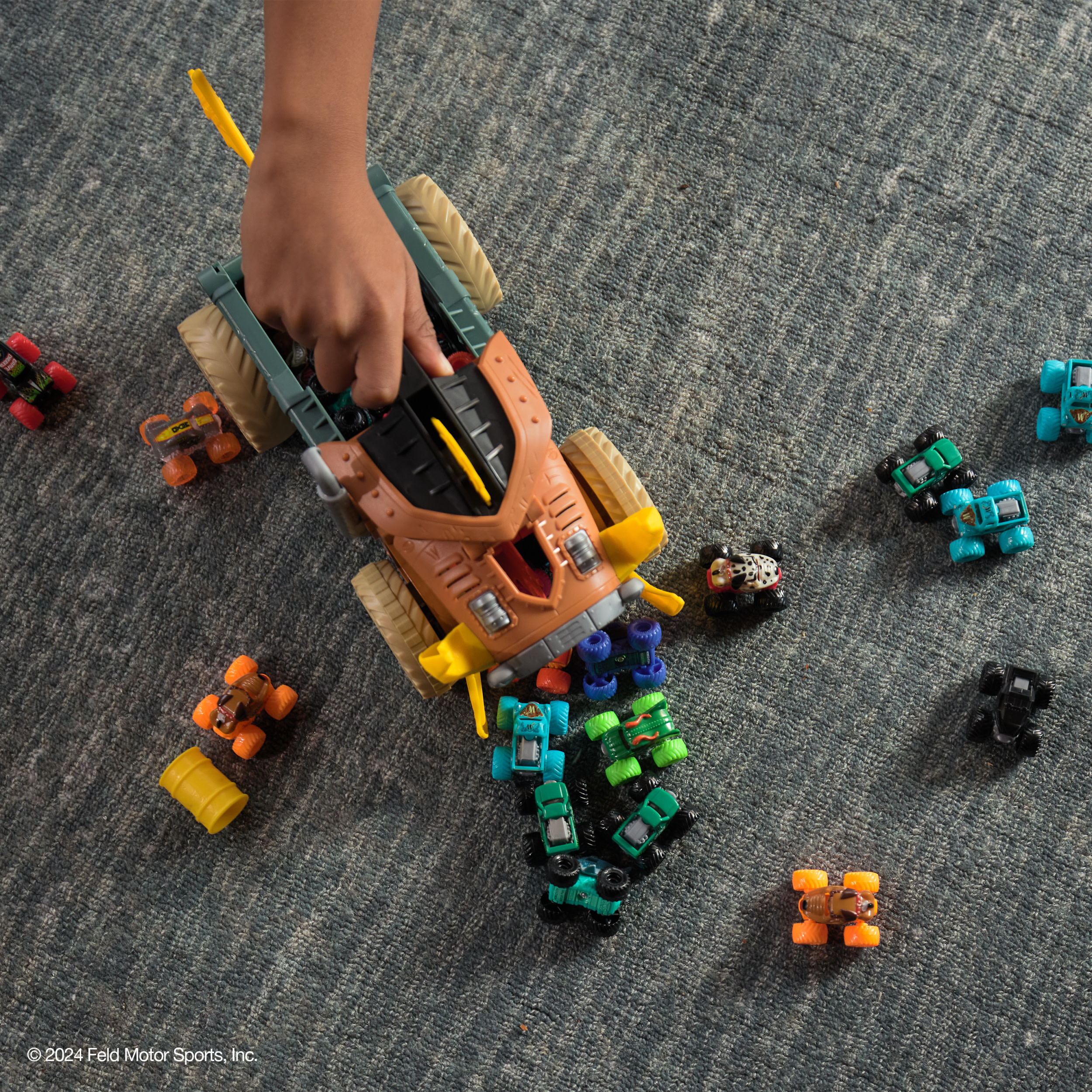 Dłoń trzymająca zabawkę ciężarówkę, otoczona rozrzuconymi miniaturowymi monster truckami na szarej wykładzinie.