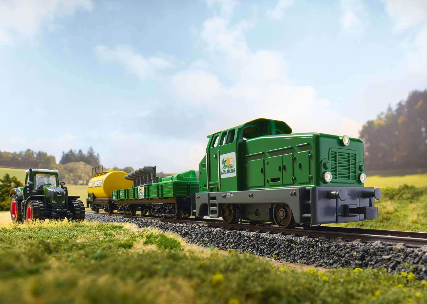 Zielony i żółty model pociągu z traktorem. Trawa i niebo w tle.