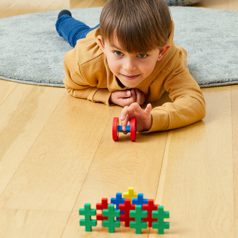 Ein Kind auf Holzboden betrachtet bunte Blöcke, wobei ein rot-blaues Spielzeug im Fokus steht.
