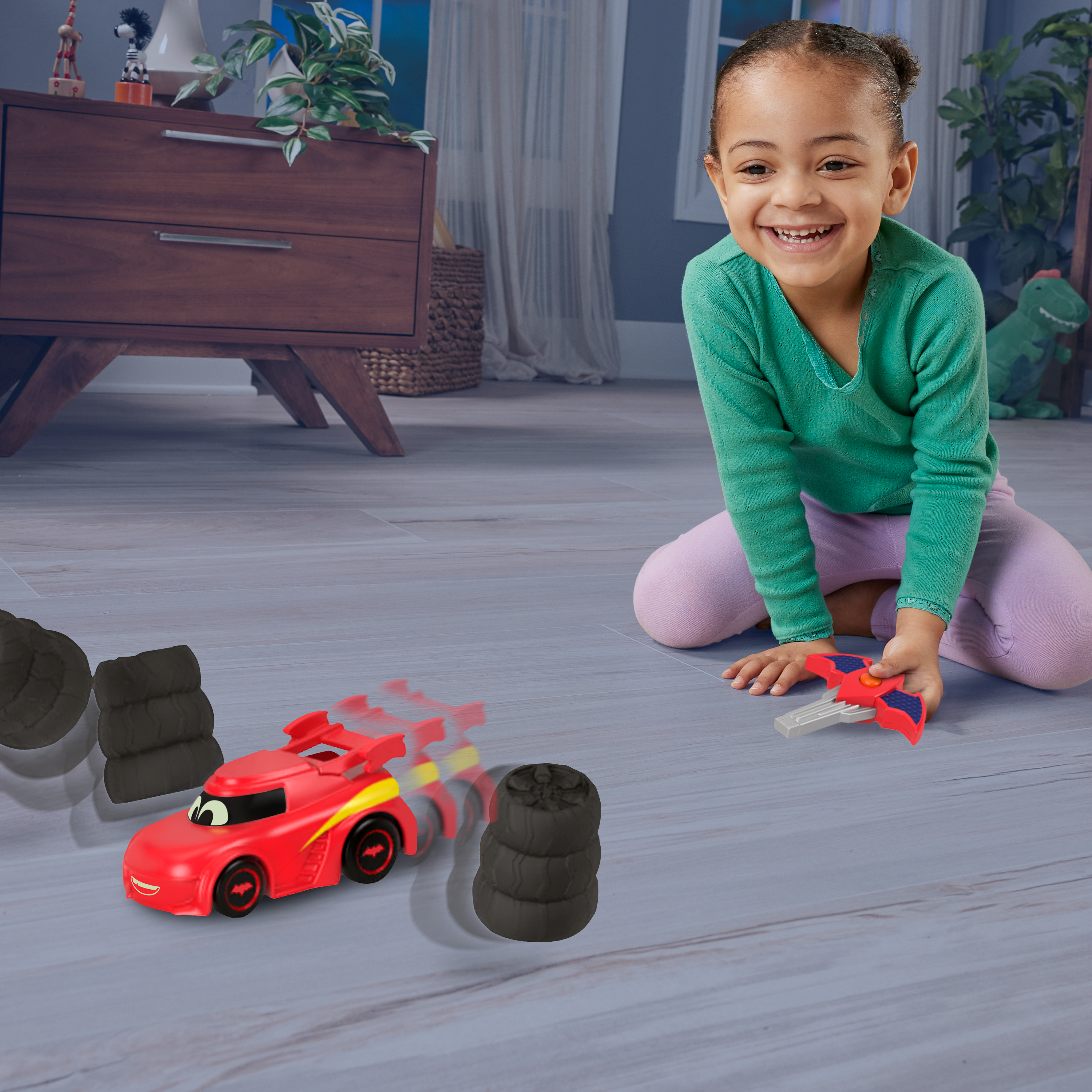 Dziewczynka bawi się czerwoną zabawką samochodem z czarnymi oponami. Obok dwa czarne stosy opon.