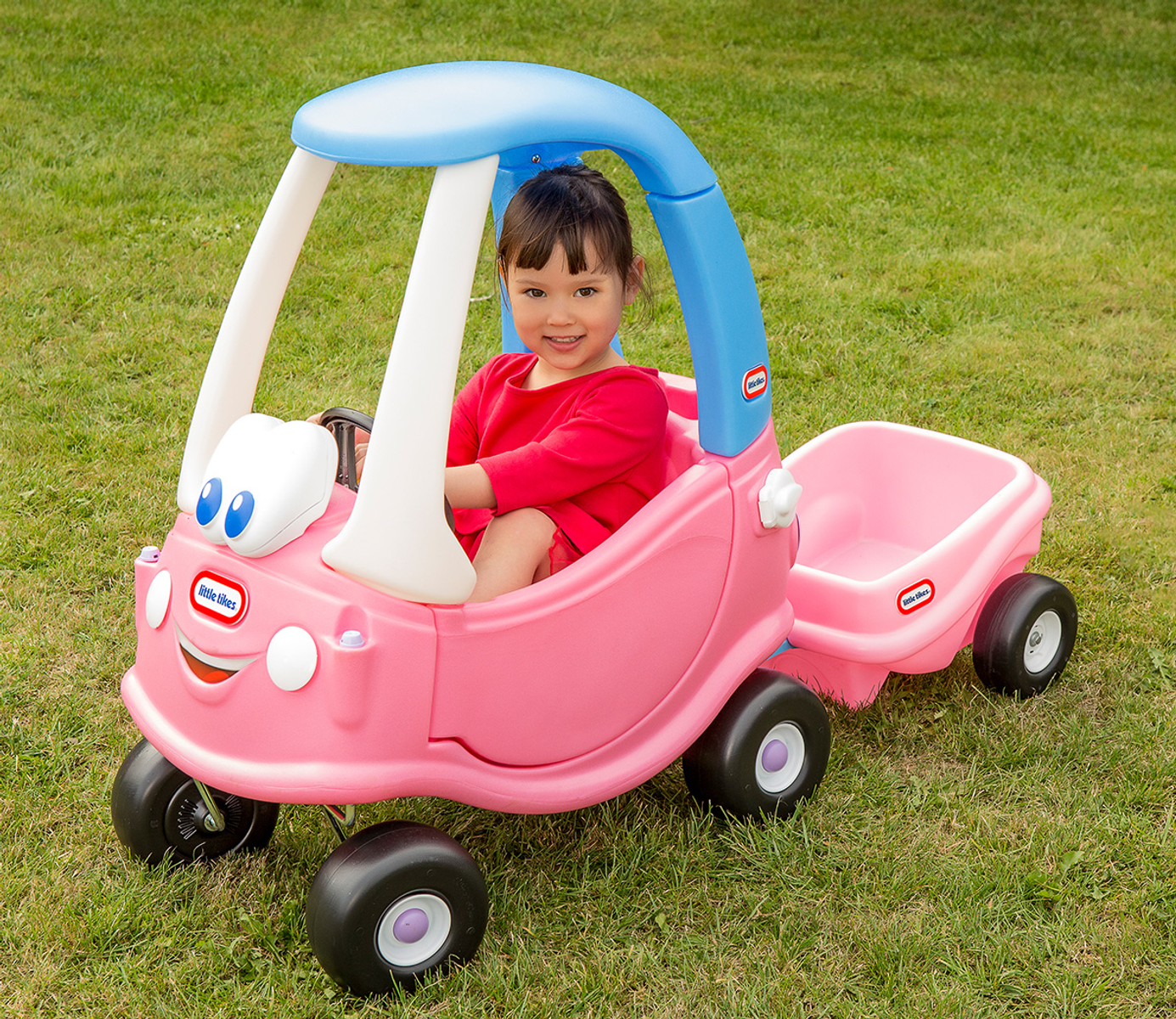 Una niña sonriendo mientras está sentada en un coche de juguete rosa con un remolque en un césped verde.