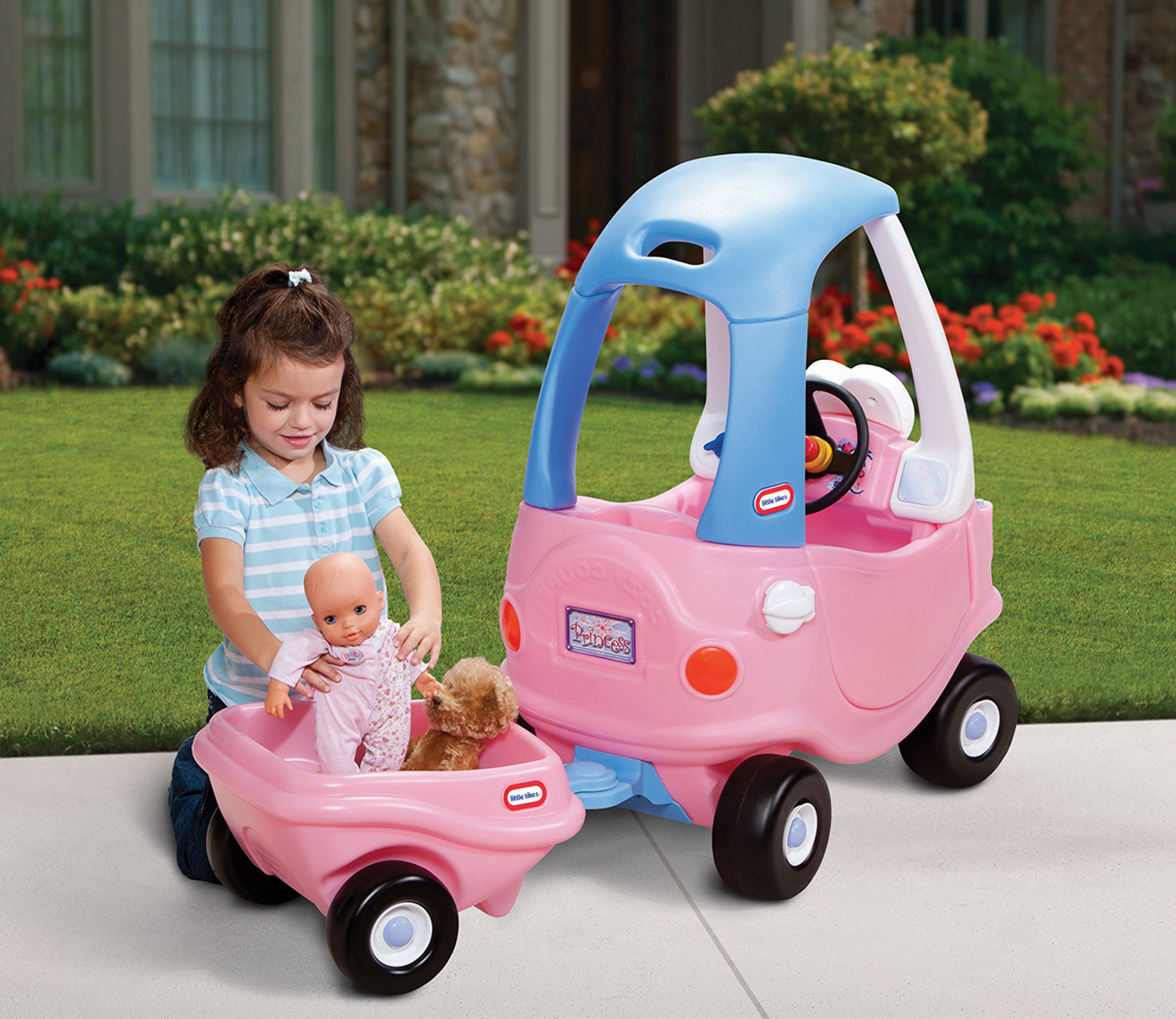 Una niña jugando con un coche de juguete rosa y un remolque rosa. Una muñeca y un osito de peluche están en el remolque.