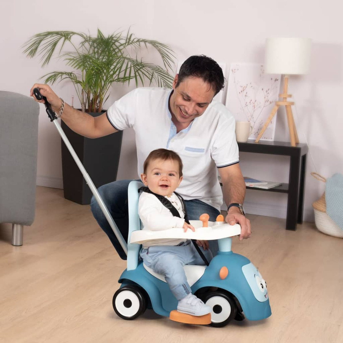 Un uomo spinge un'auto giocattolo blu con un bambino seduto all'interno. Sono in una stanza con mobili.