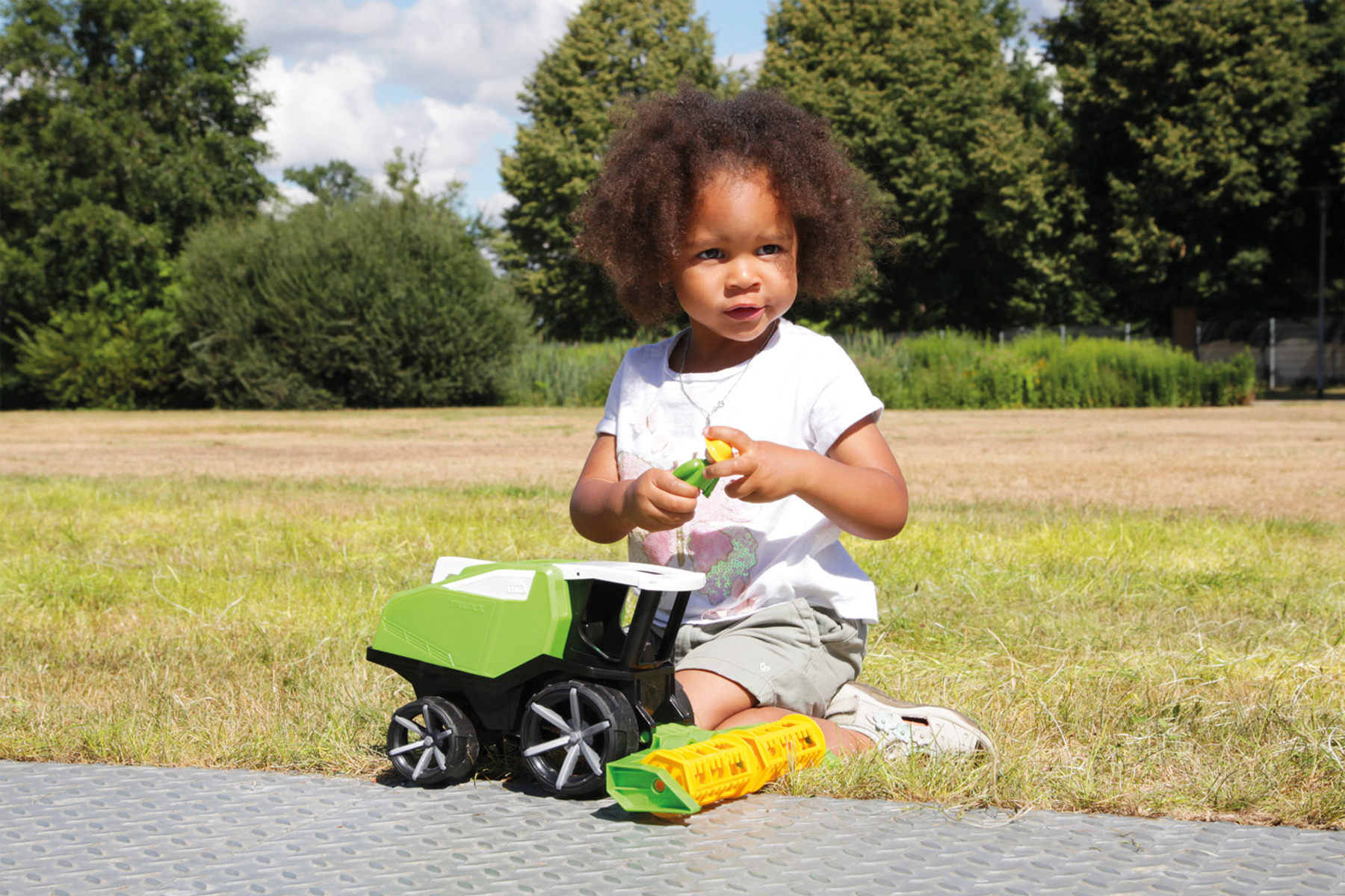 Niña sentada en la hierba jugando con un tractor de juguete verde y amarillo. Día soleado, árboles verdes al fondo.