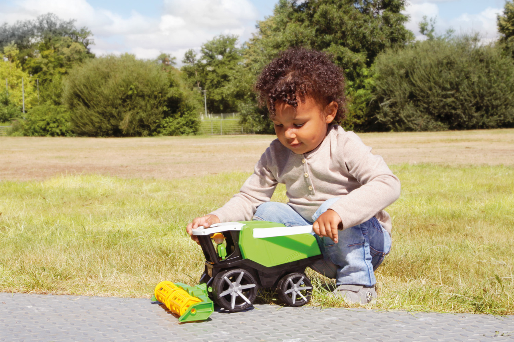 Niña jugando con un tractor de juguete verde y amarillo. Arrodillada sobre una superficie gris, hierba y árboles al fondo.
