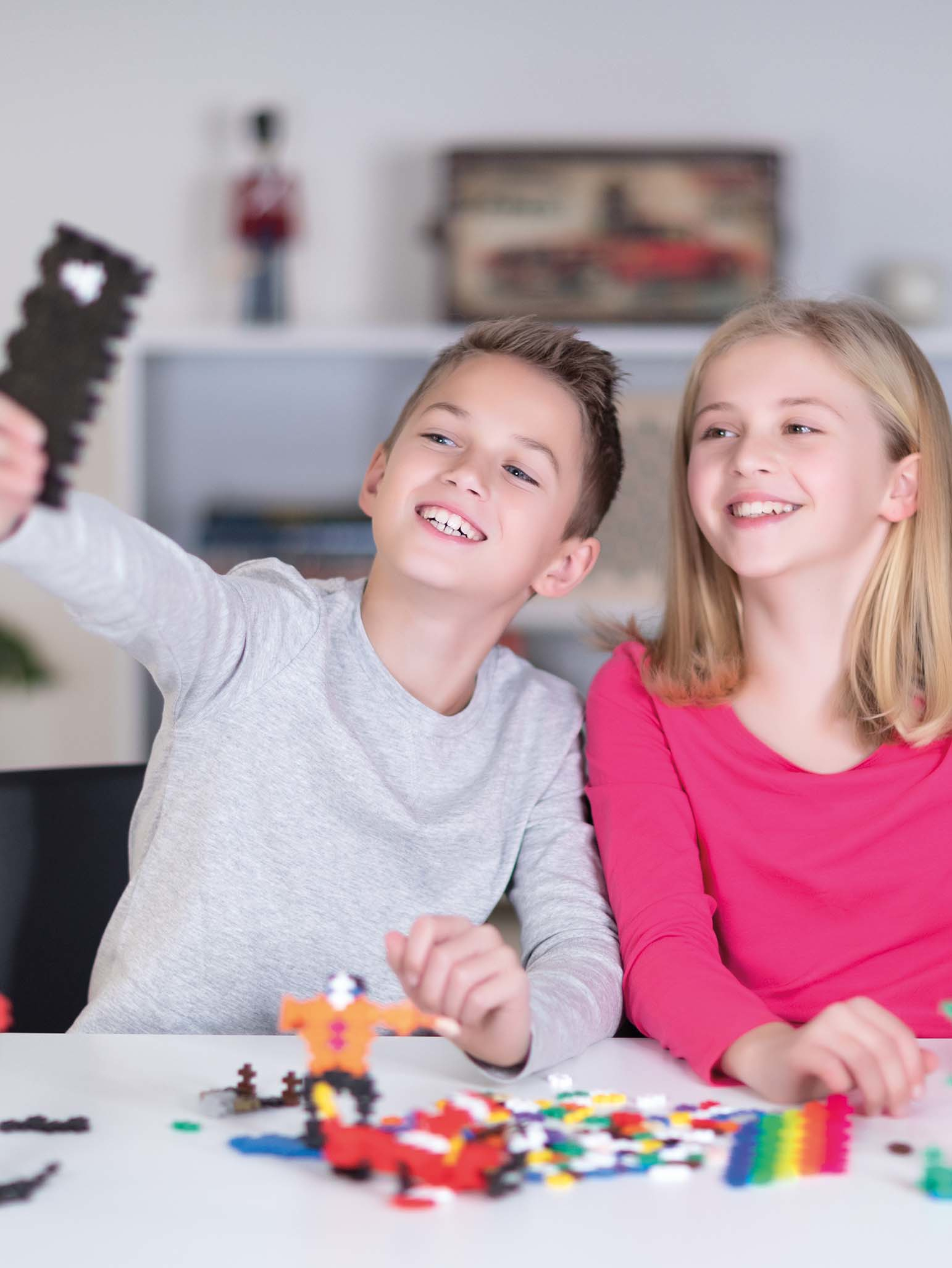 Zwei Kinder machen ein Selfie mit einem Spielzeug. Junge grau, Mädchen pink. Spielzeugblöcke auf dem Tisch.