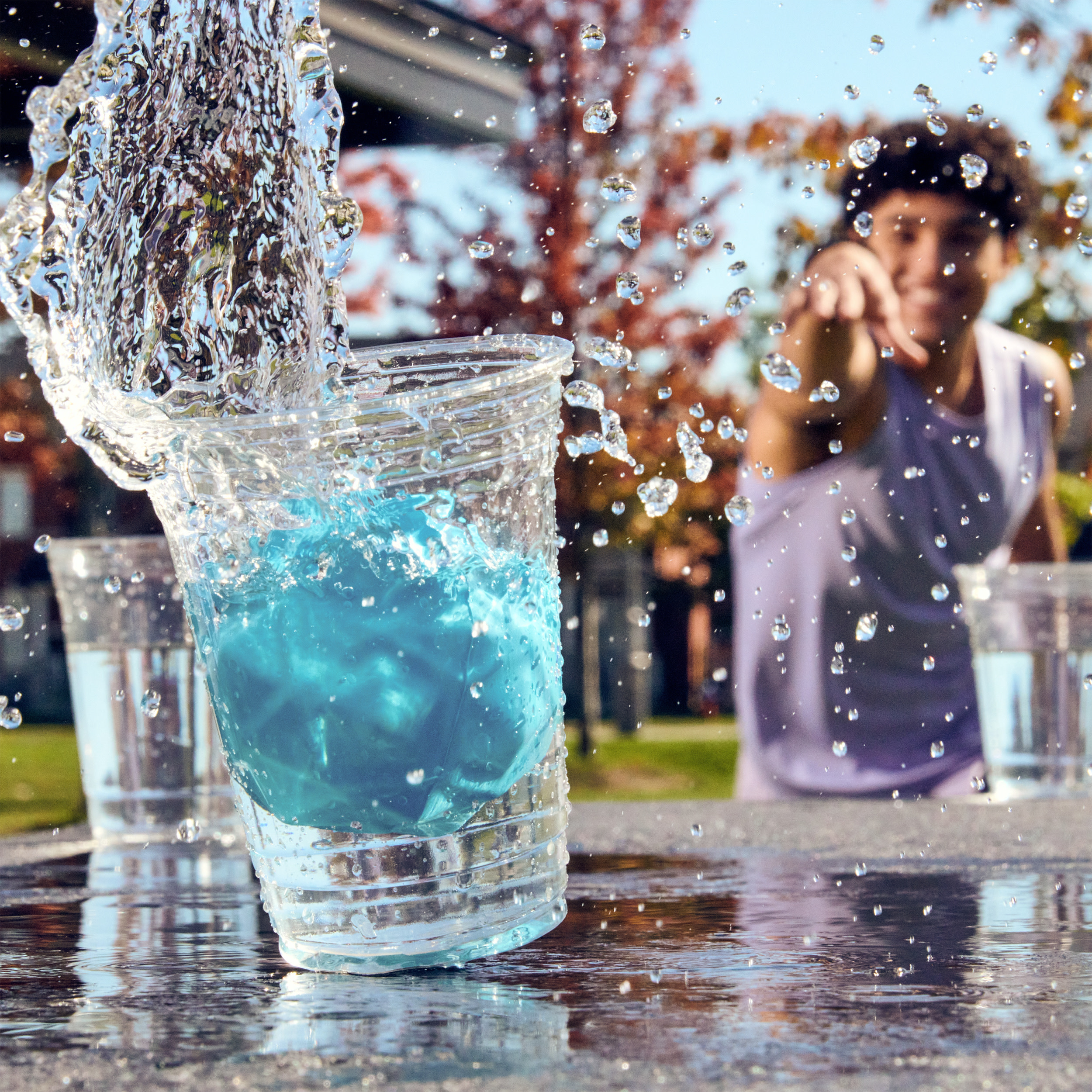 Woda rozpryskuje się ze szklanki wypełnionej niebieskim płynem. Osoba wskazuje na rozmyte tło.