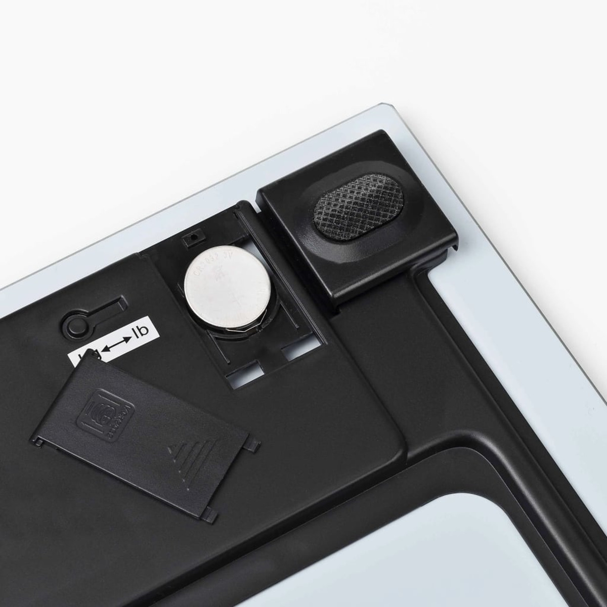 Le dessous d'une balance est montré, avec le compartiment de la batterie ouvert et une pile ronde. L'étiquette de l'unité indique lb.
