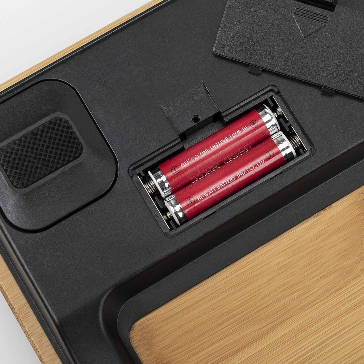 Spód wagi pokazujący komorę baterii. Dwie czerwone baterie w środku. Czarna podstawa i drewno.