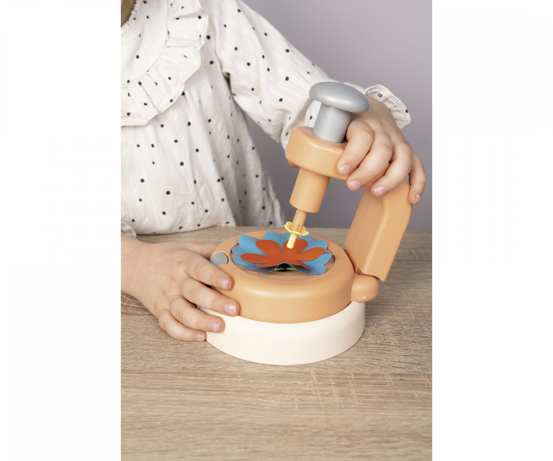 Dziecko używa maszyny do rękodzieła, naciskając na przedmiot w kształcie kwiatu. Maszyna jest beżowo-biała.