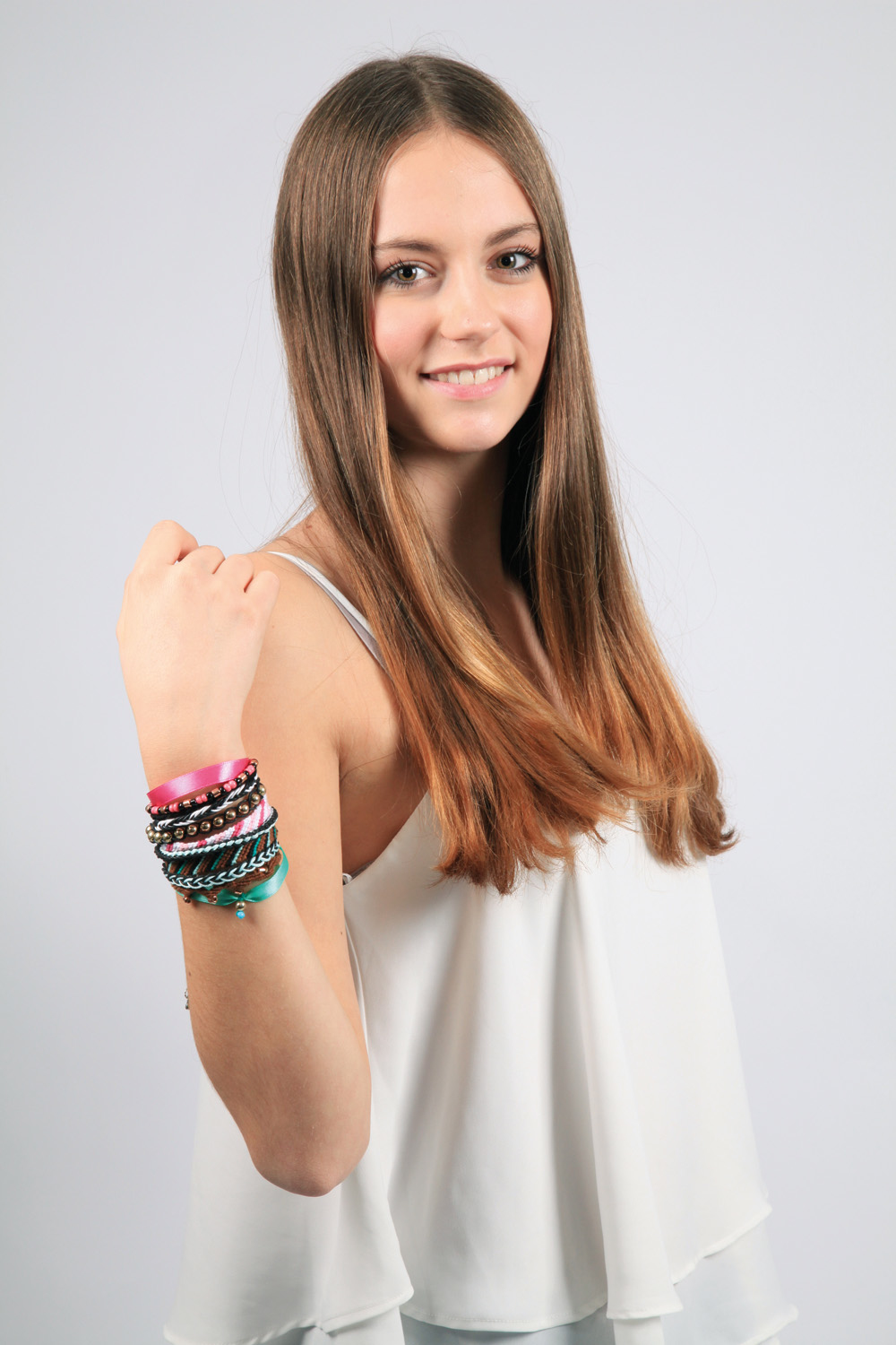Młoda kobieta z długimi brązowymi włosami ma na sobie biały top i bransoletki na lewym ramieniu. Uśmiecha się.
