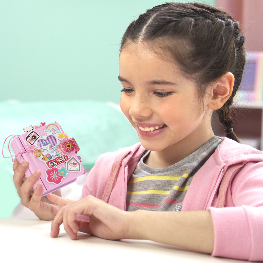 Uśmiechnięta dziewczyna trzyma różowy pamiętnik z naklejkami. Ma na sobie różową kurtkę i pasiastą koszulę.