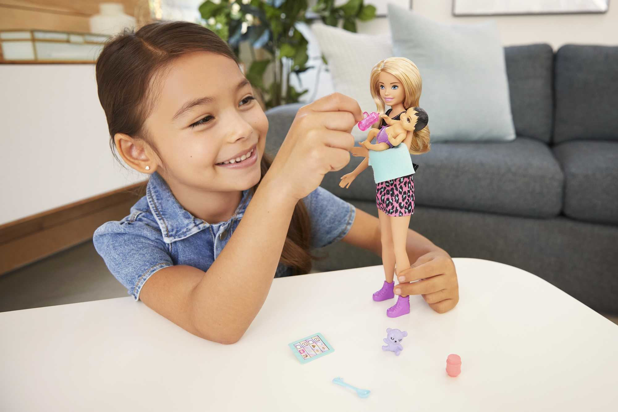 Ein Mädchen spielt mit einer Barbie-Puppe, die eine Babypuppe hält. Zubehör wie Löffel und Teddybär sind vorhanden.