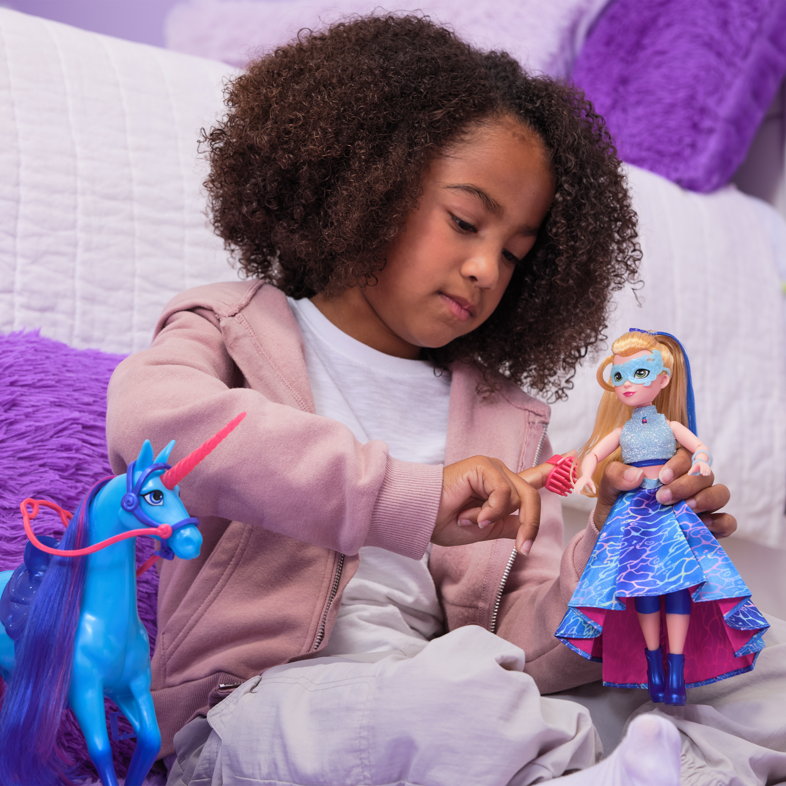 Una niña sosteniendo una muñeca con un unicornio azul. La muñeca tiene una máscara y un vestido azul. Cama de fondo.