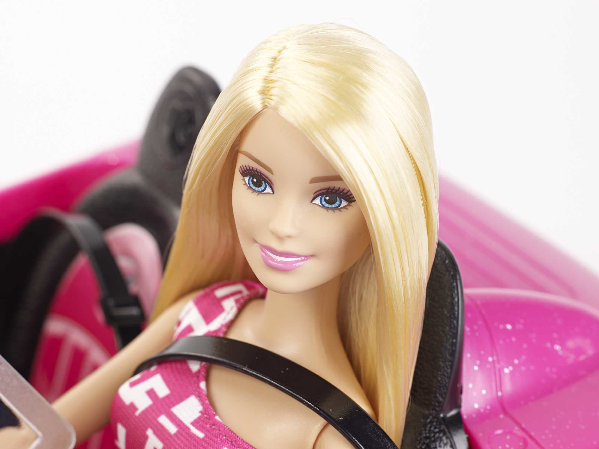 Blond Barbie, uśmiechnięta, patrząca w kamerę, w różowym samochodzie.