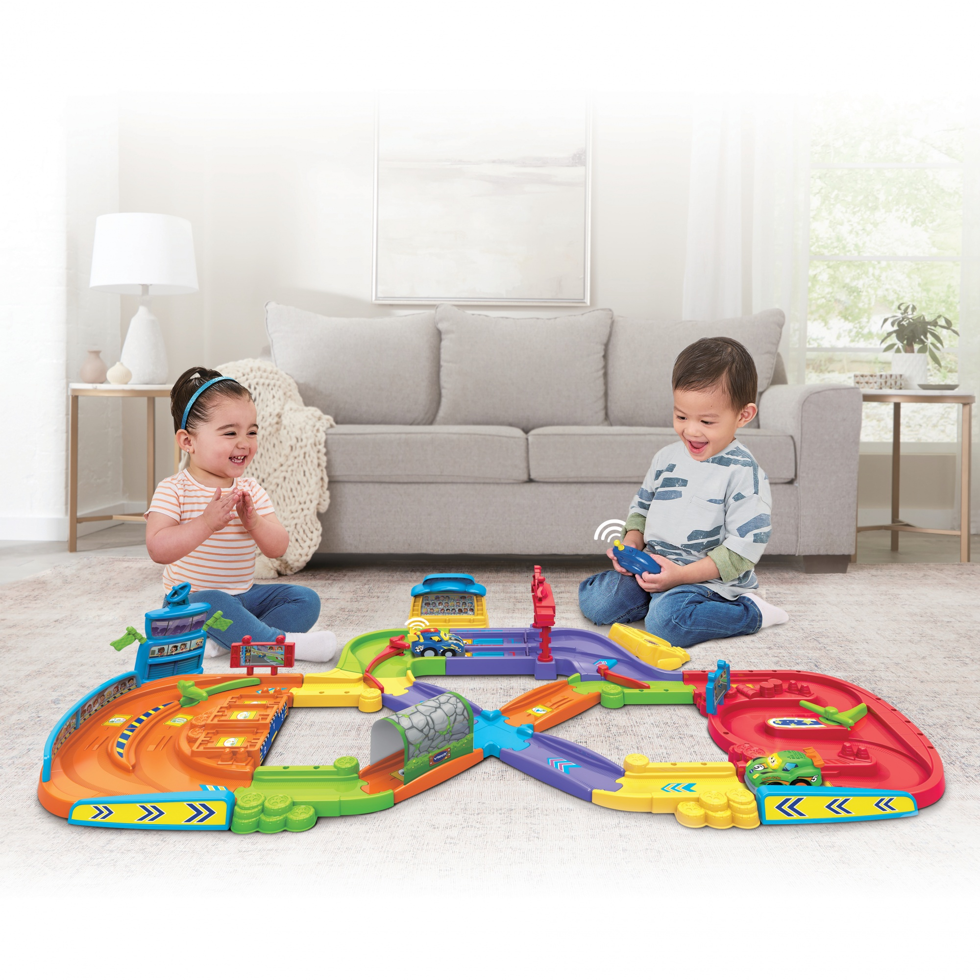 Dwoje dzieci bawiących się kolorowym torem wyścigowym zabawek, z sofą w tle.