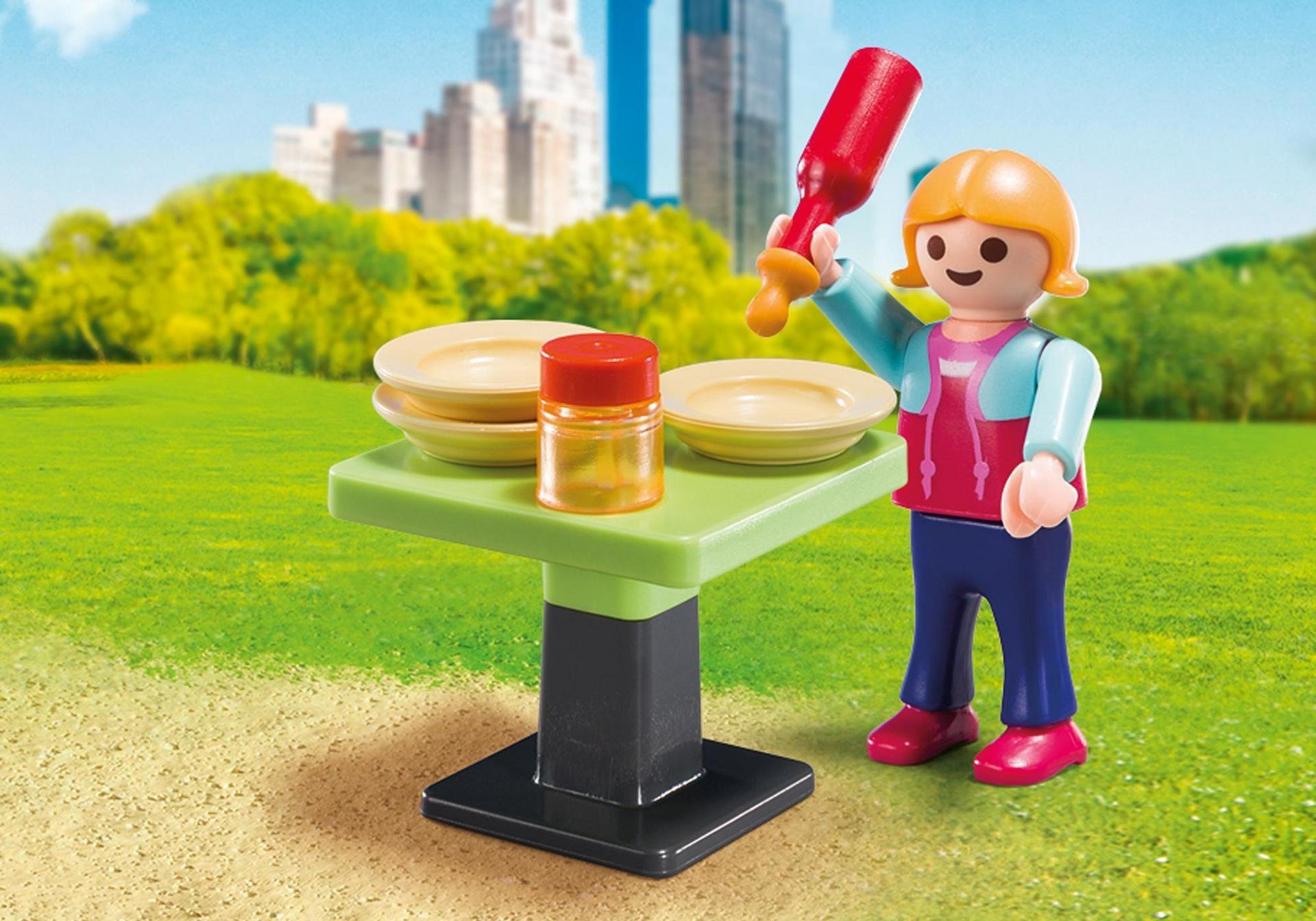 Eine Playmobil-Figur mit einer Flasche in der Nähe eines Tisches mit Tellern und einem Glas, in einem Park.
