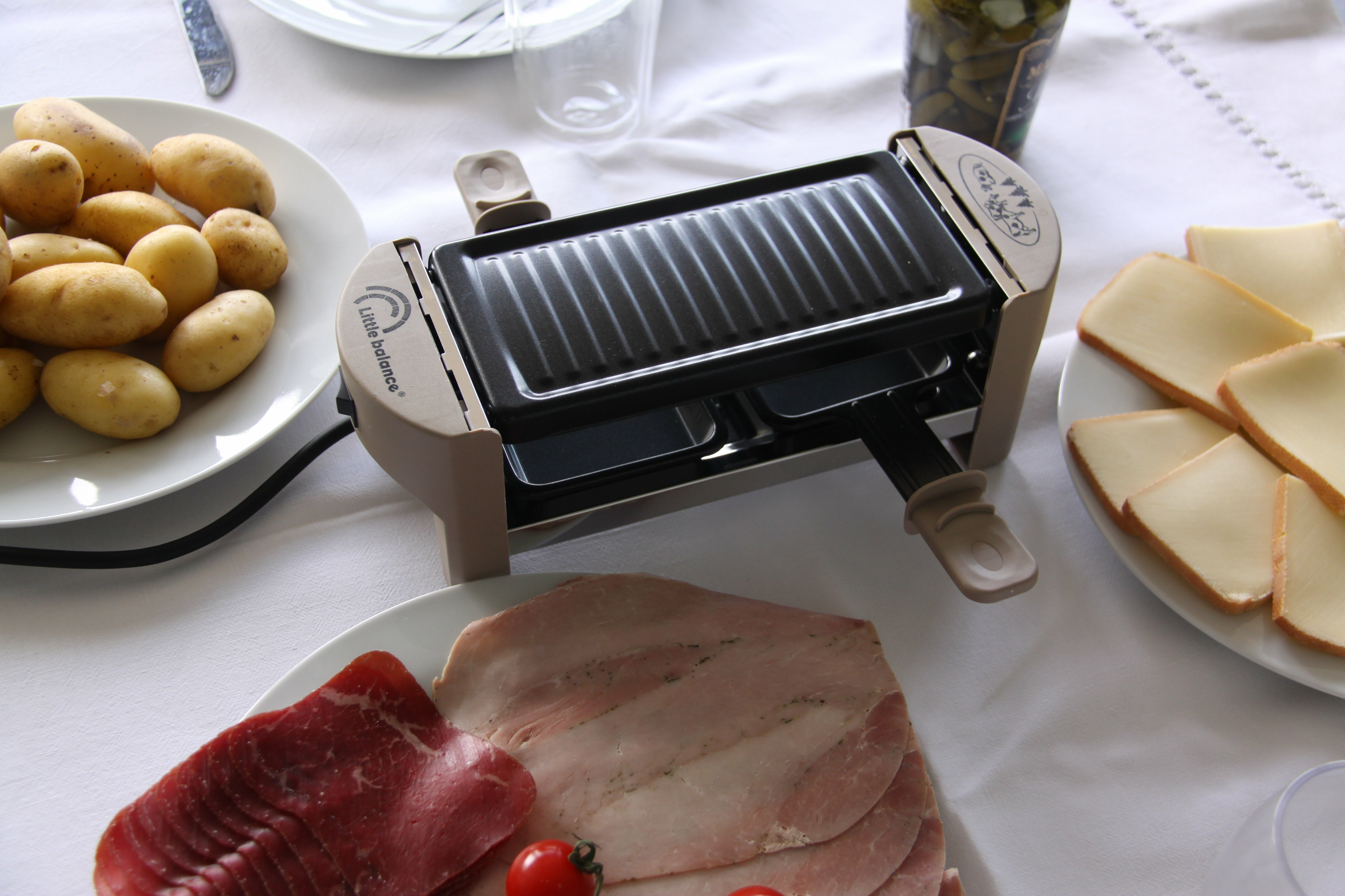 Stół z grillem do raclette, ziemniakami, serem i szynką, przygotowany do posiłku.