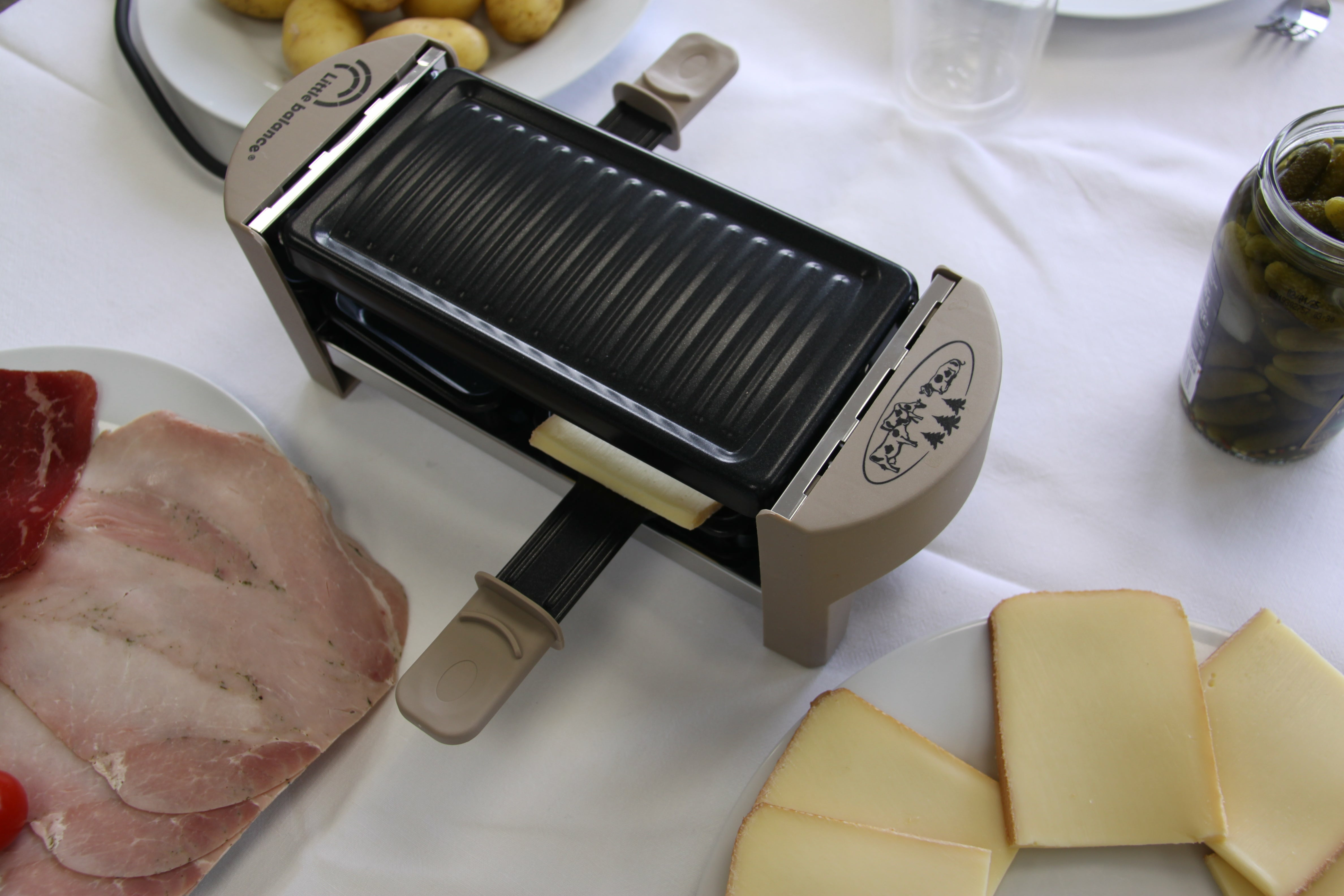 Grill do raclette z jedzeniem, takim jak ser, ziemniaki i mięso, na białym stole.