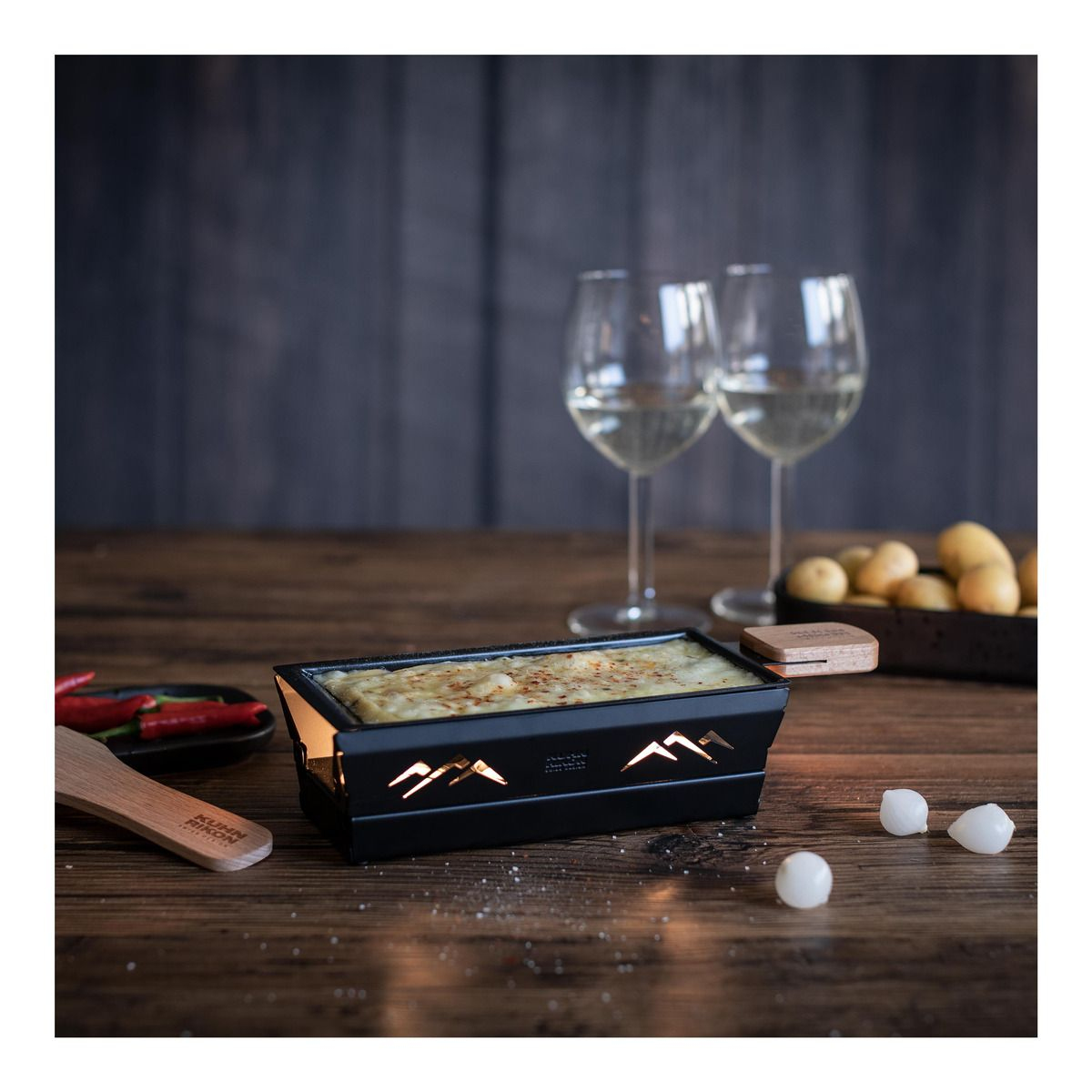 Raclette-Grill mit geschmolzenem Käse. Zwei Weingläser. Holztisch.