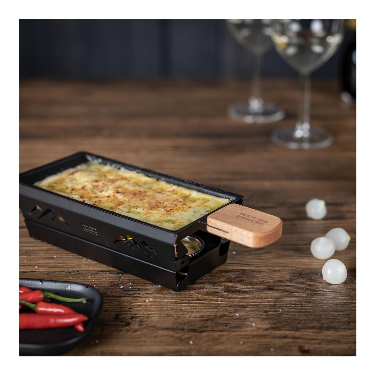 Raclette-Grill mit geschmolzenem Käse. Holztisch. Rote Chilischoten.