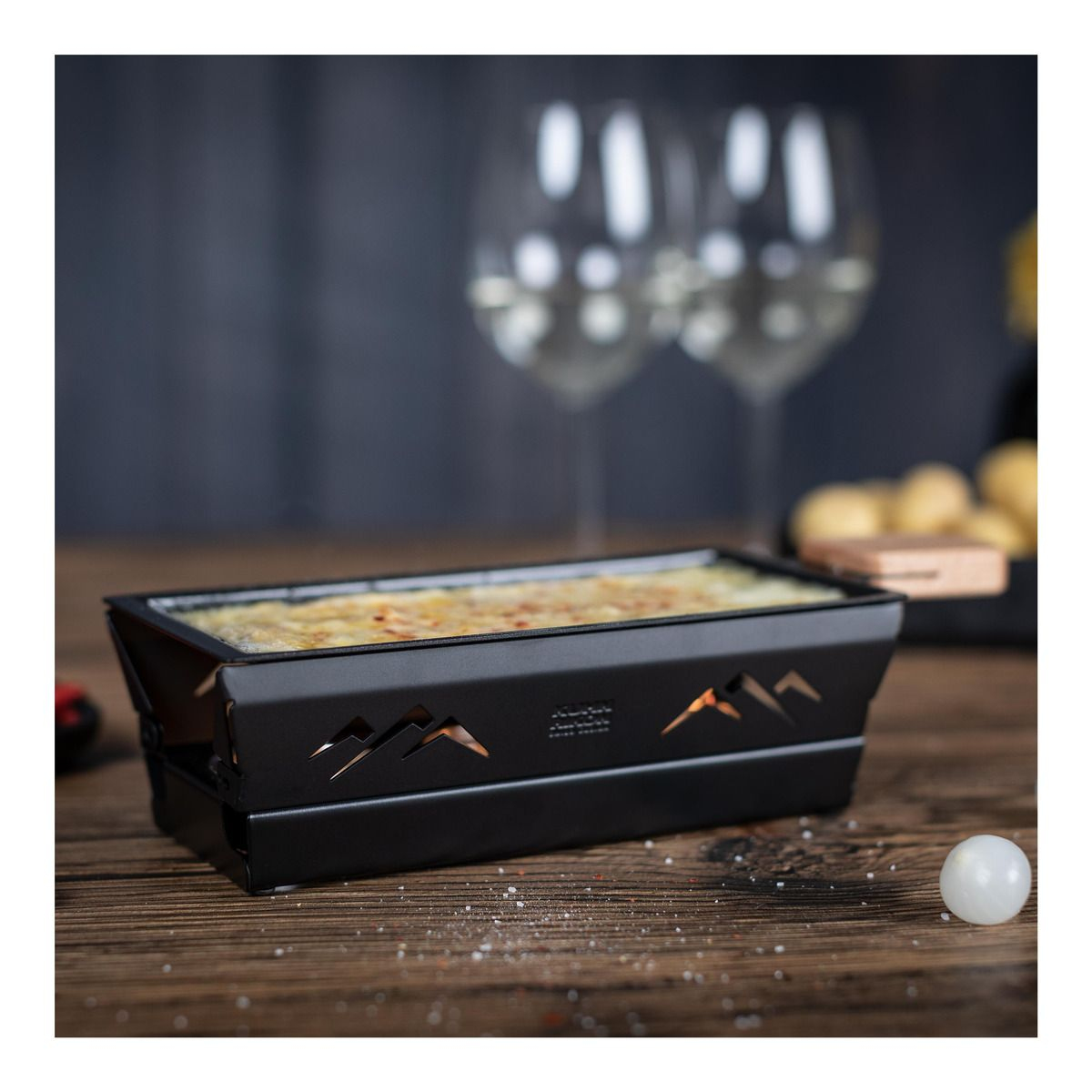Schwarzer Tisch-Raclette-Grill mit Käse und Weingläsern. Dunkler Hintergrund. Weiße Oberfläche.