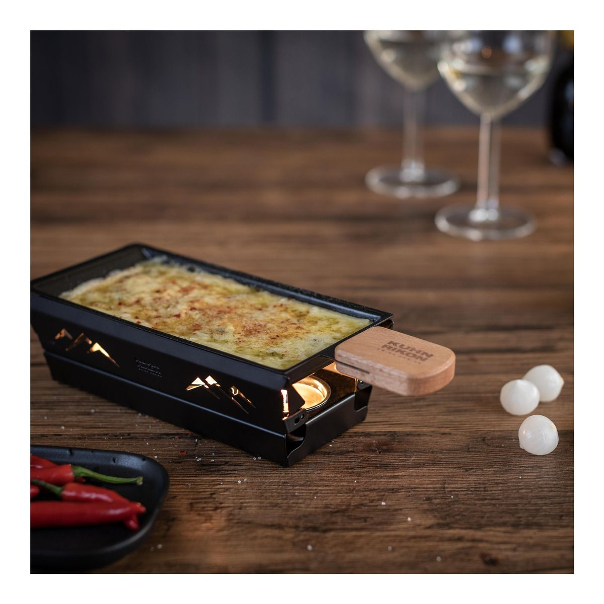 Raclette-Grill mit geschmolzenem Käse. Holztisch. Rote Chilischoten.