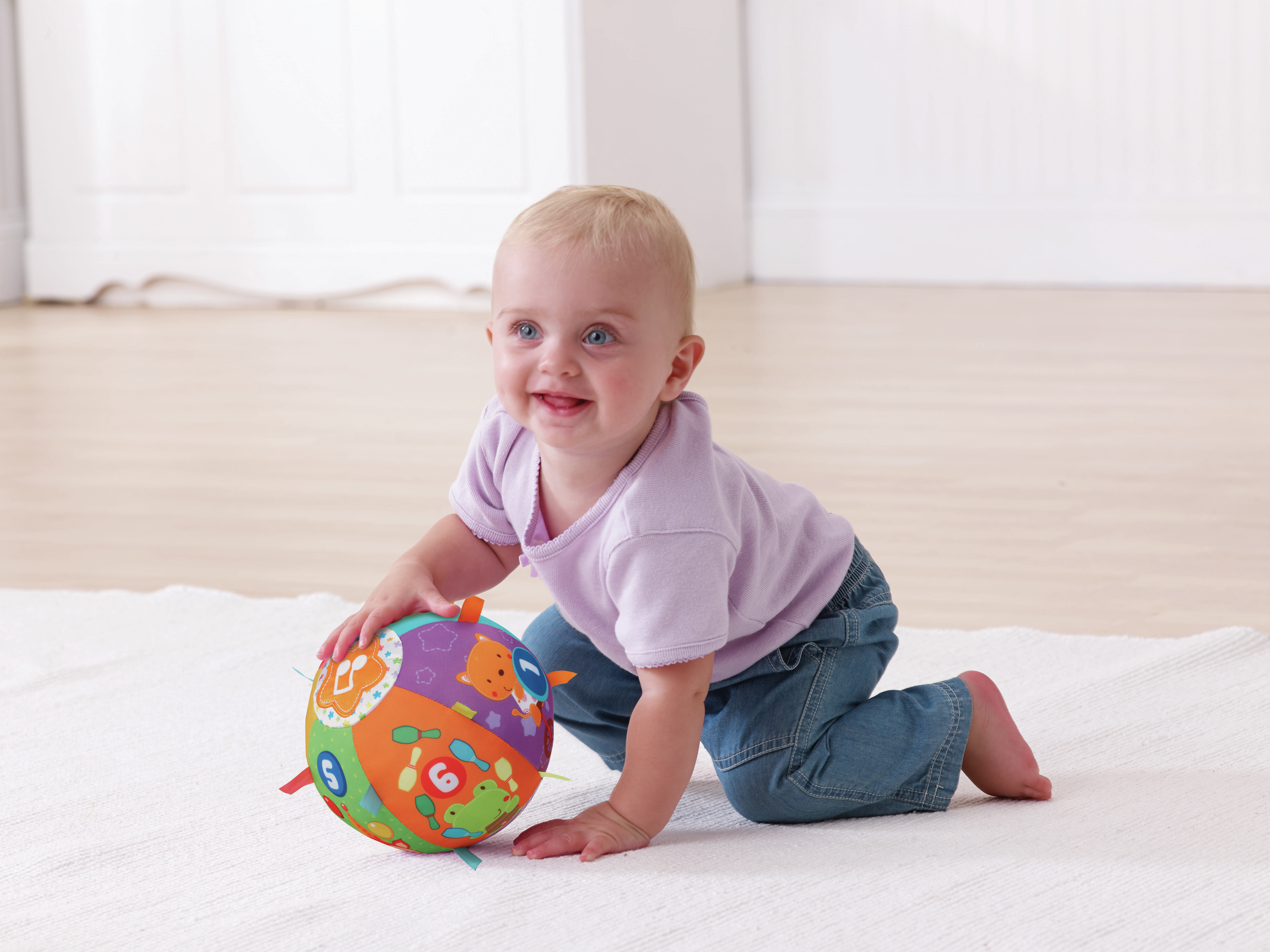 Uśmiechnięte dziecko czołga się w stronę kolorowej piłki na białym dywanie.