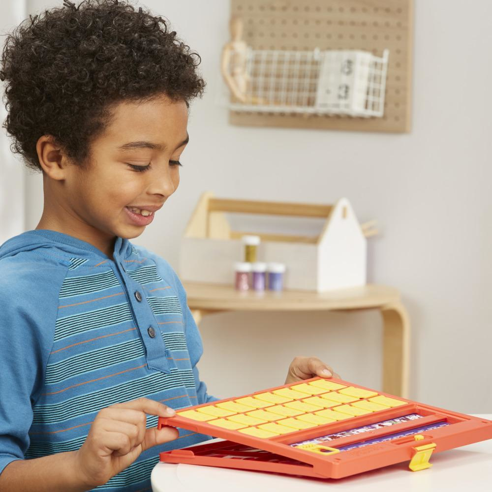 Jongen opent een Guess Who-spel. Oranje bord met gele kaarten. Een witte tafel en blauwe achtergrond.