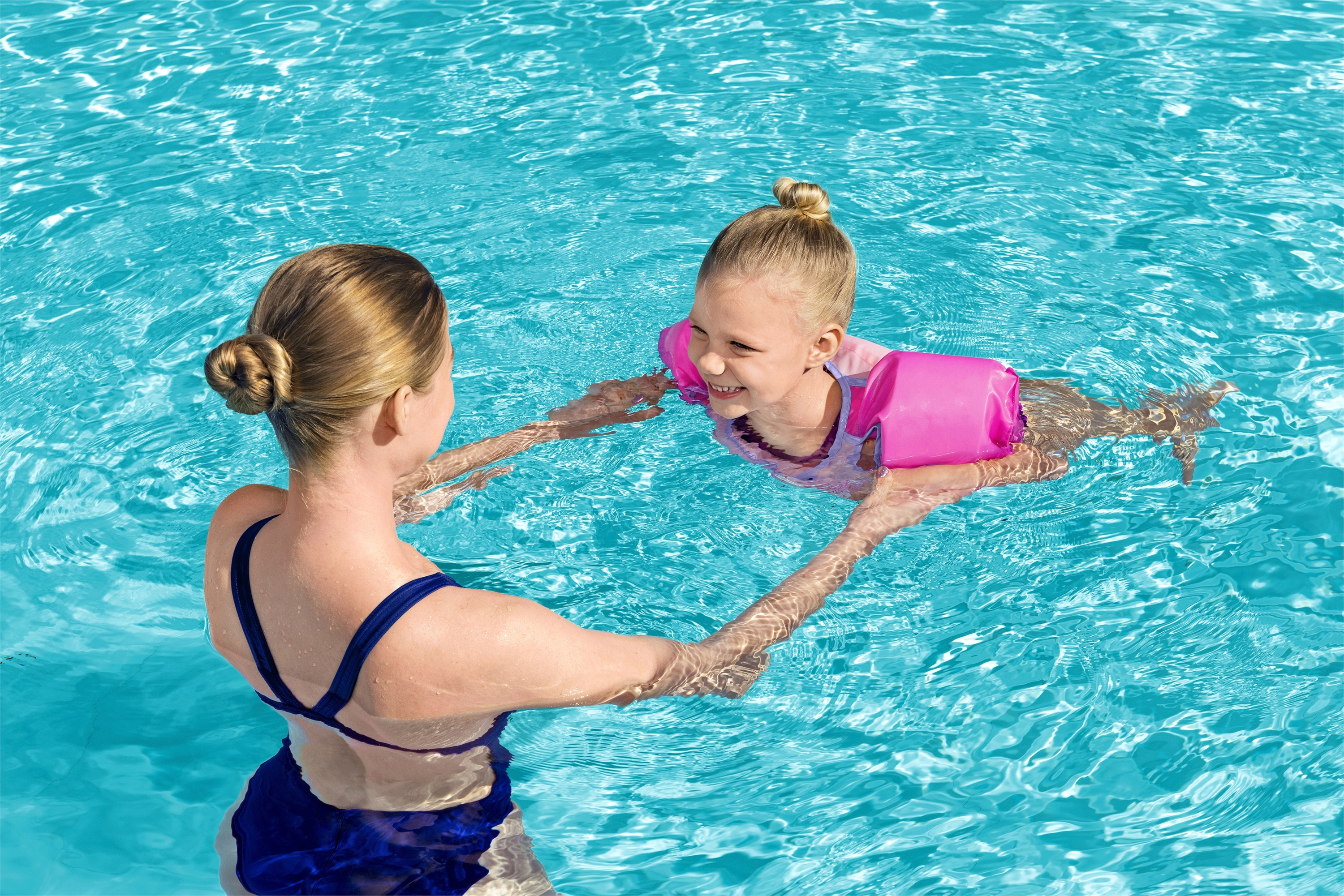 Kobieta trzymająca małą dziewczynkę w basenie, obie się uśmiechają, w strojach kąpielowych i rękawkach.