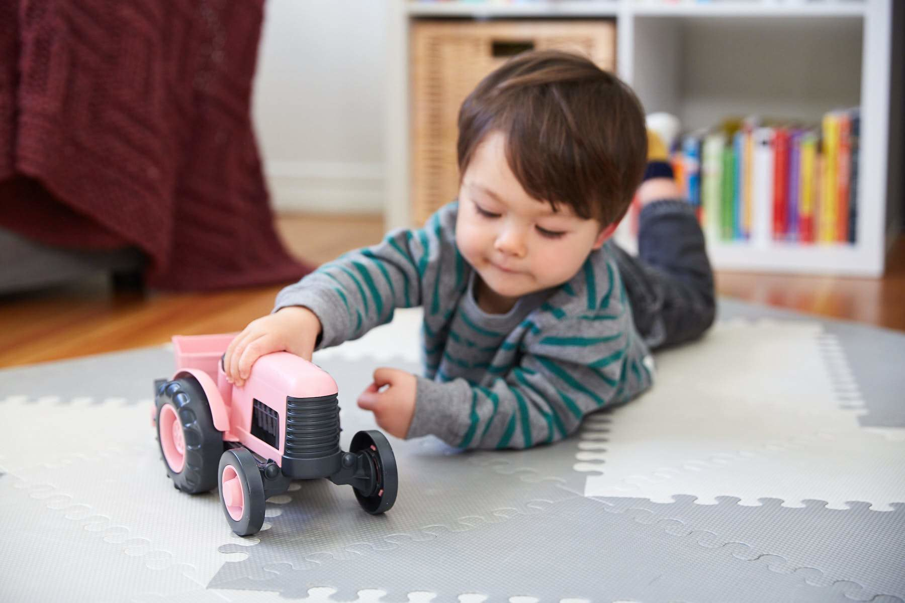 Małe dziecko bawiące się różowym traktorem na szarej i białej macie do zabawy przed półką na książki.