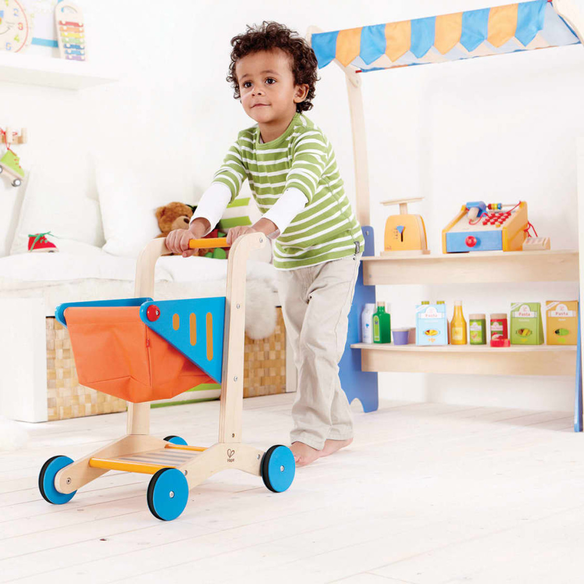 Un niño empuja un carrito de compras de madera en un juego de tienda. El fondo tiene estantes y un mostrador.