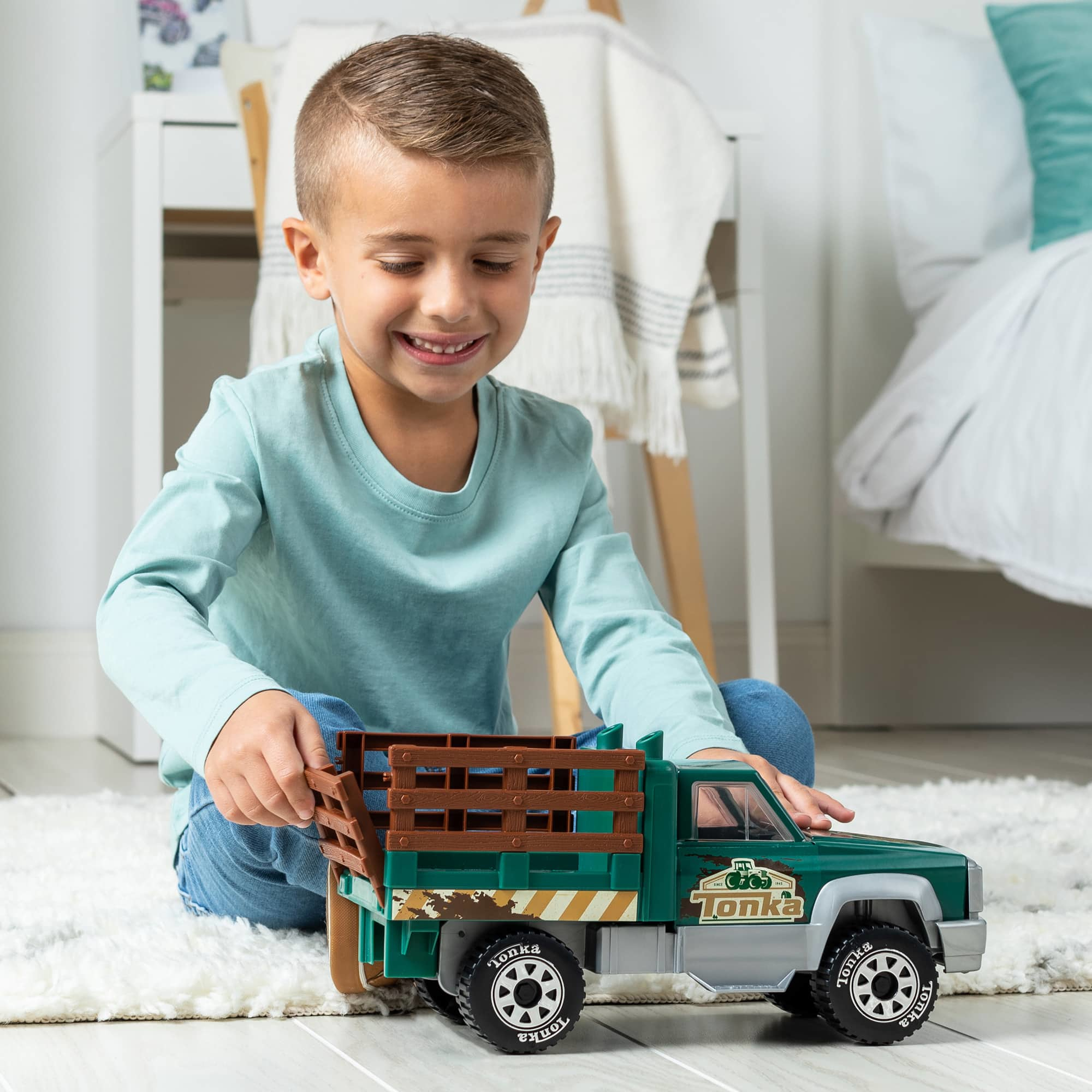 Ein Junge spielt mit einem grün-grauen Spielzeug-LKW. Der LKW steht auf einem weißen Teppich in einem Raum.
