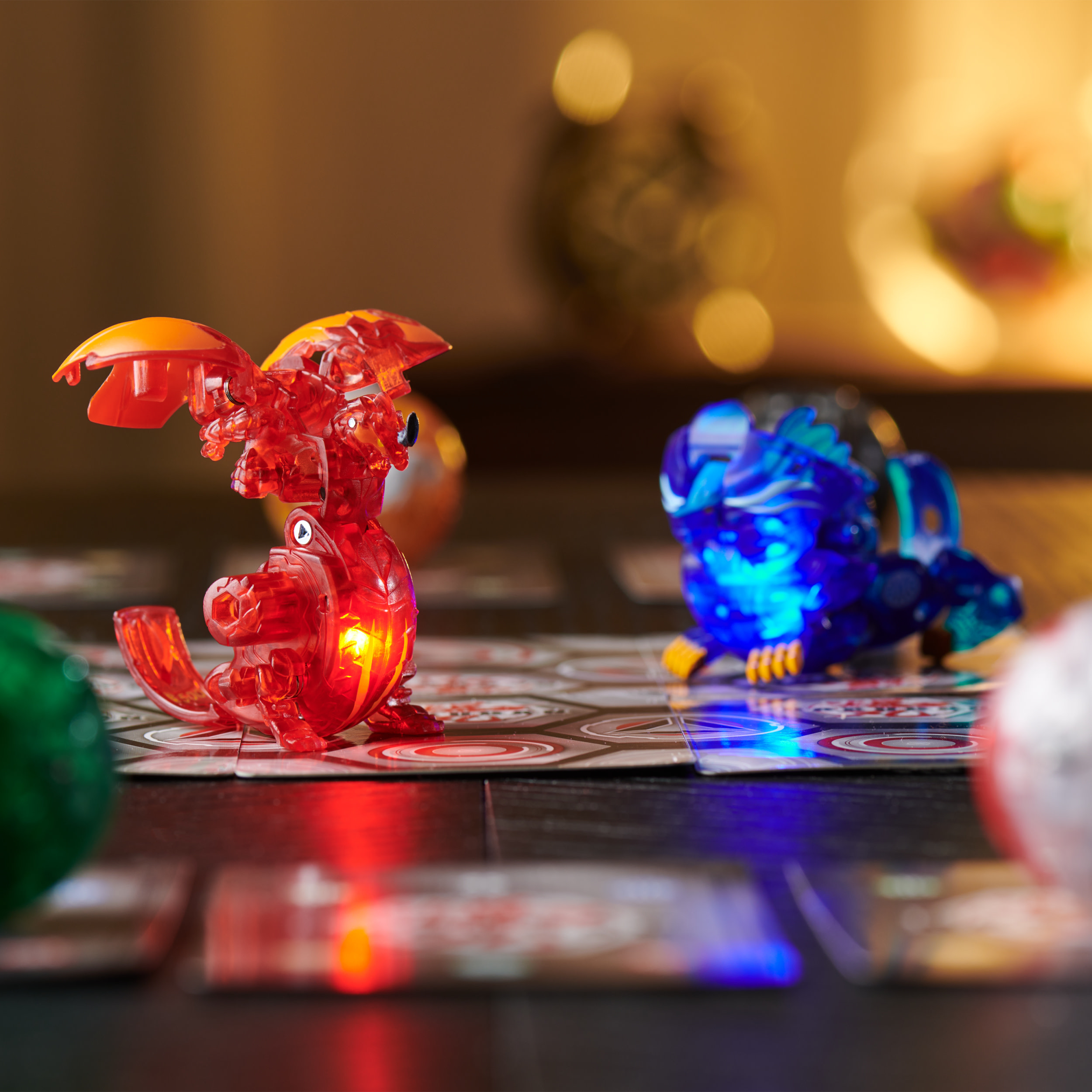 Dos figuras brillantes de Bakugan en una alfombra de juego con cartas, iluminadas en un entorno oscuro.