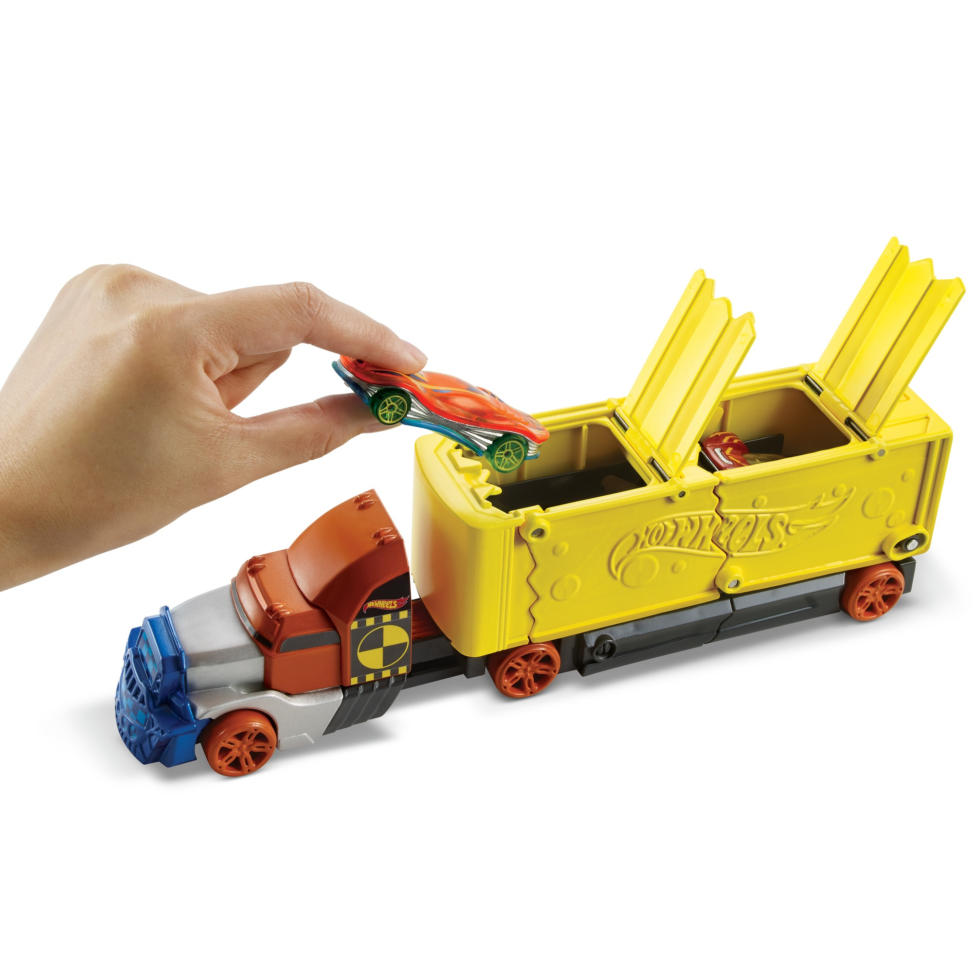 Ręka umieszcza zabawkowy samochód w ciężarówce Hot Wheels. Ciężarówka jest żółta, pomarańczowa i niebieska.