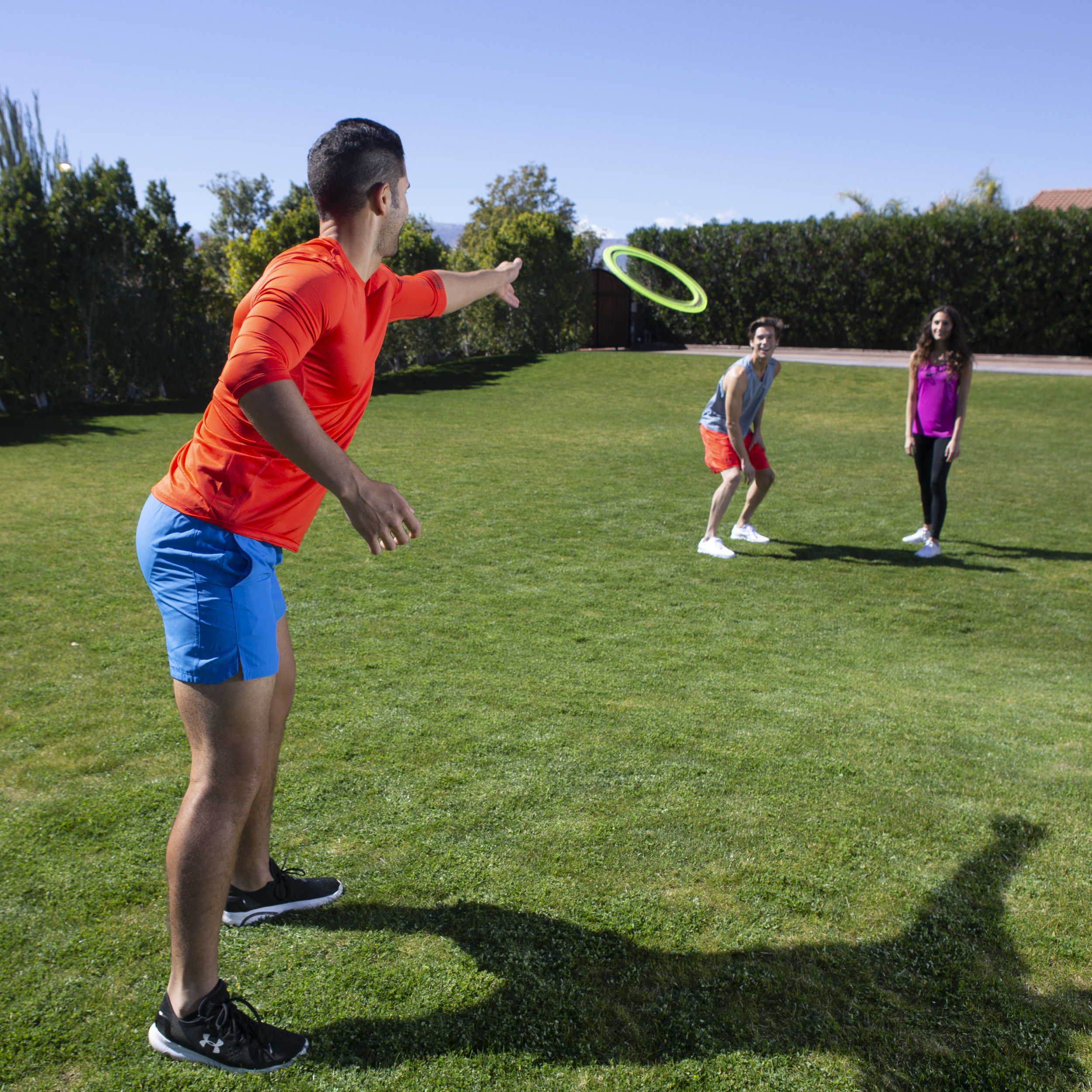Trzy osoby grają w frisbee na zielonym trawniku pod błękitnym niebem. Jeden rzuca, dwóch patrzy.