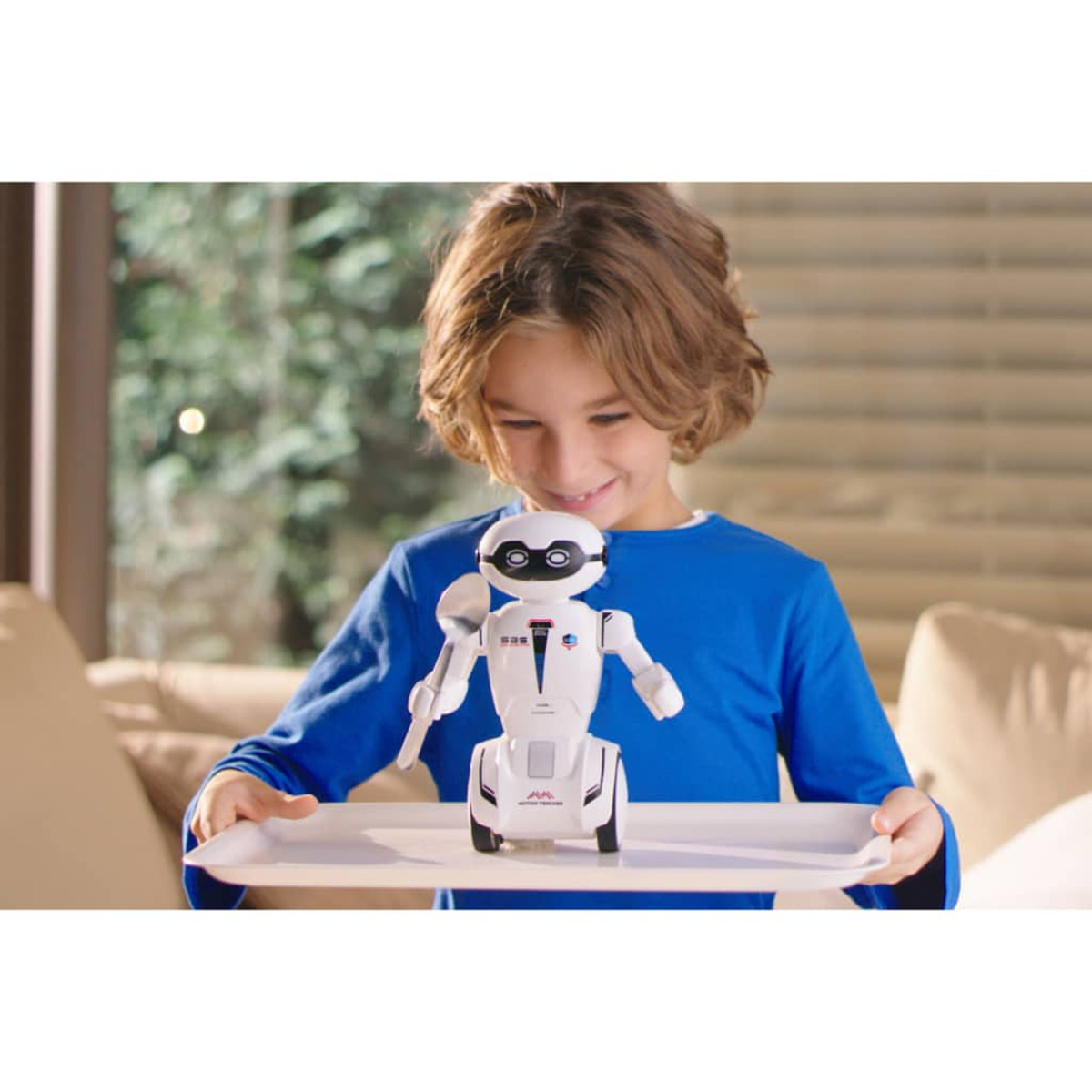 Chłopiec trzyma zabawkę robota na tacy. Robot jest biały. Tło pokazuje sofę.