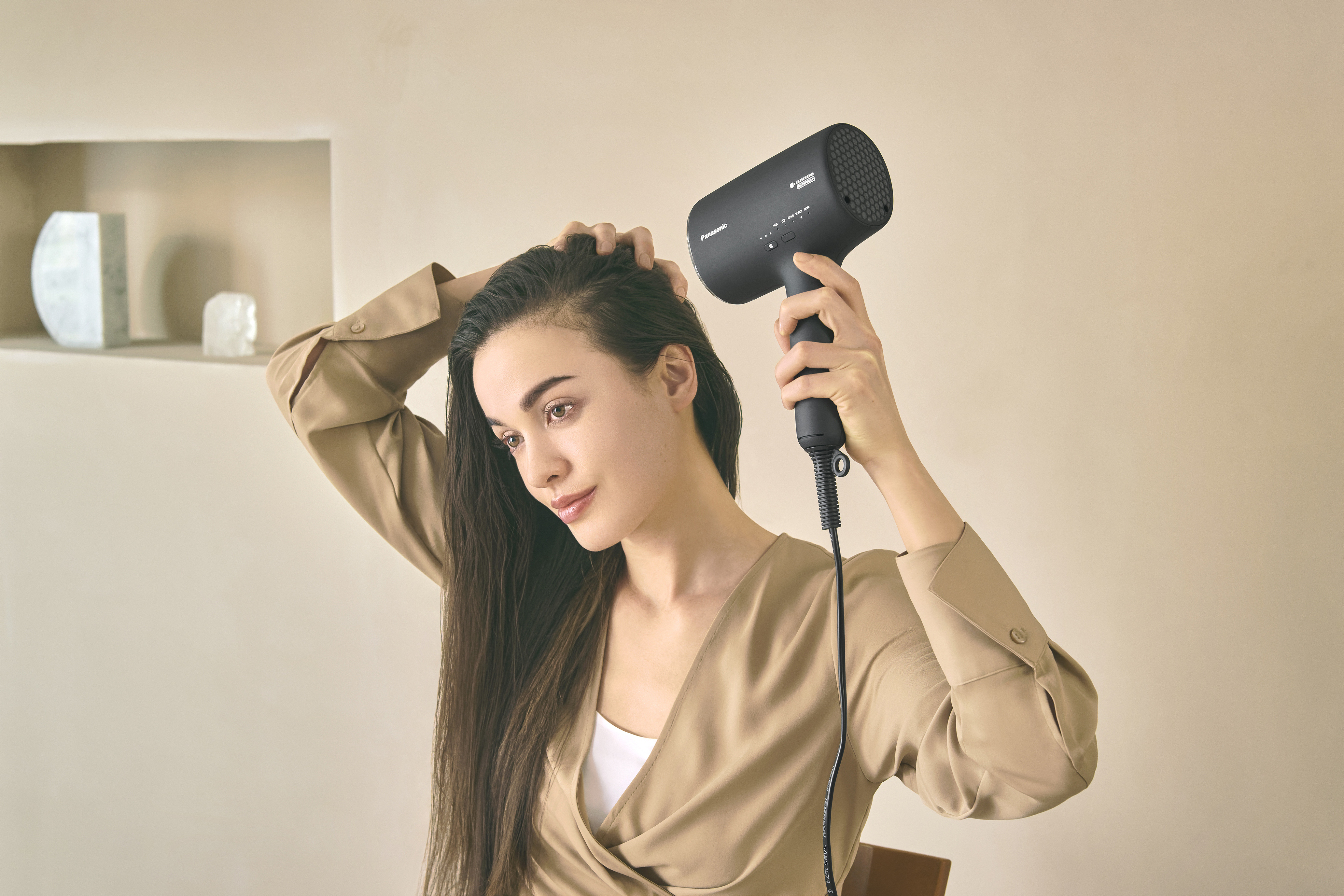 Kobieta używająca czarnej suszarki do włosów, scena w pomieszczeniu, beżowe tło.
