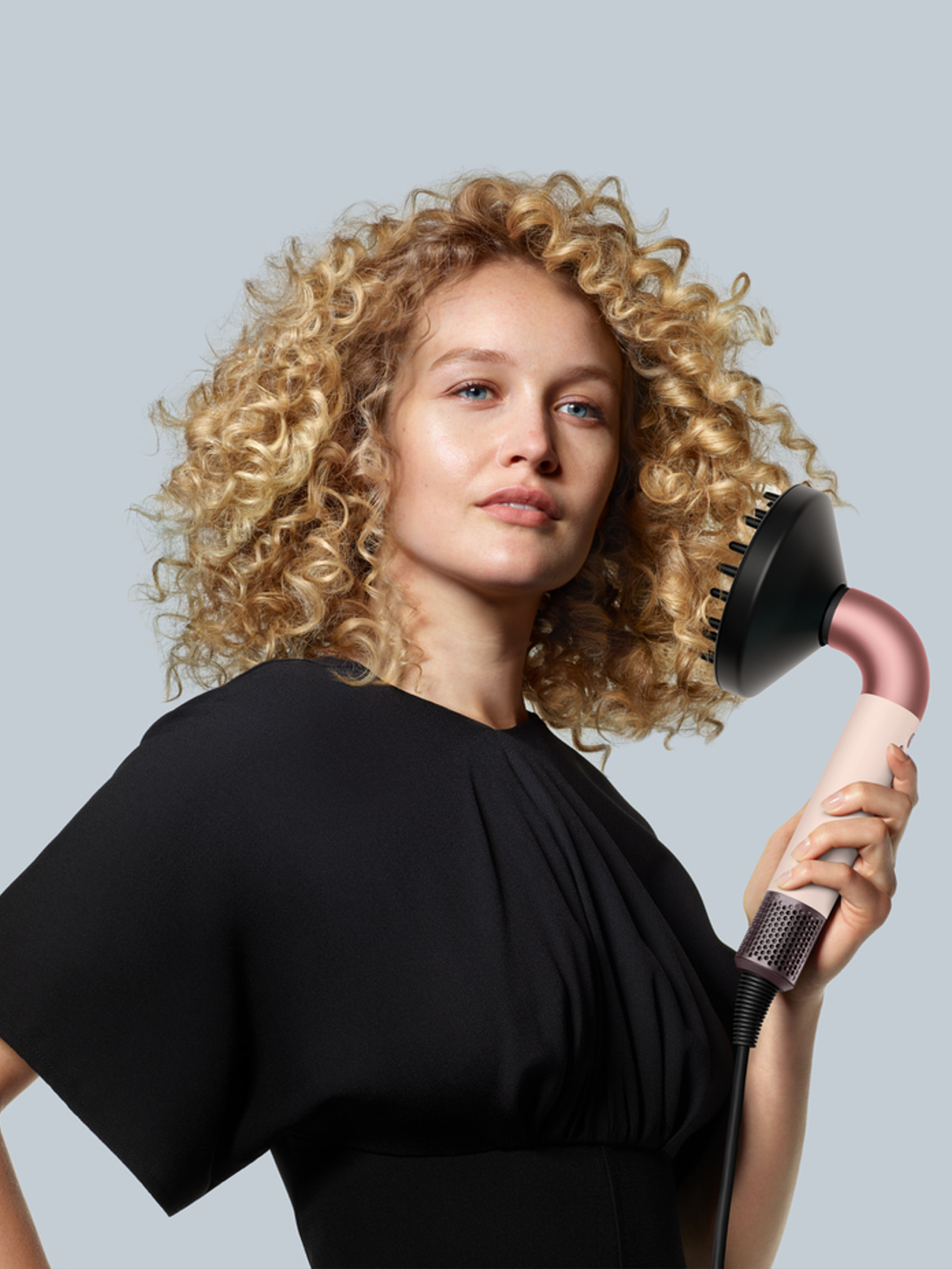 Eine Frau mit lockigem blondem Haar hält einen Fön mit Diffusor. Der Hintergrund ist hellblau.