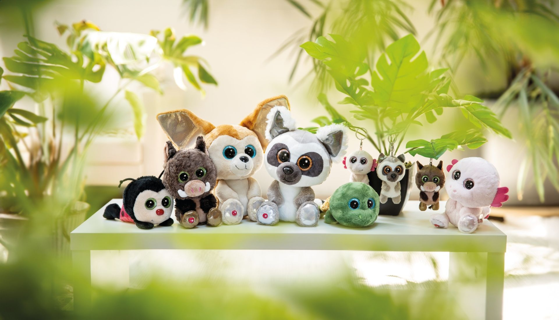 Grupa pluszowych zabawek na białej powierzchni. Zielone rośliny w tle. Słoneczna scena.