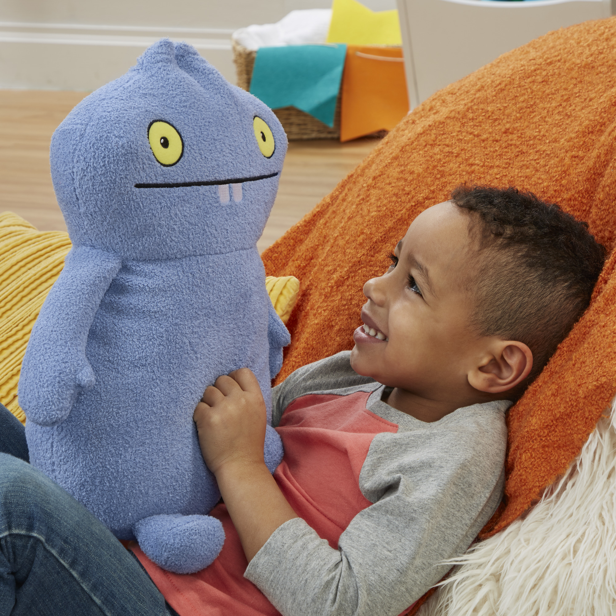 Ein Kind lächelt ein blaues Ugly Dolls Plüschtier an. Oranges Sofa im Hintergrund.