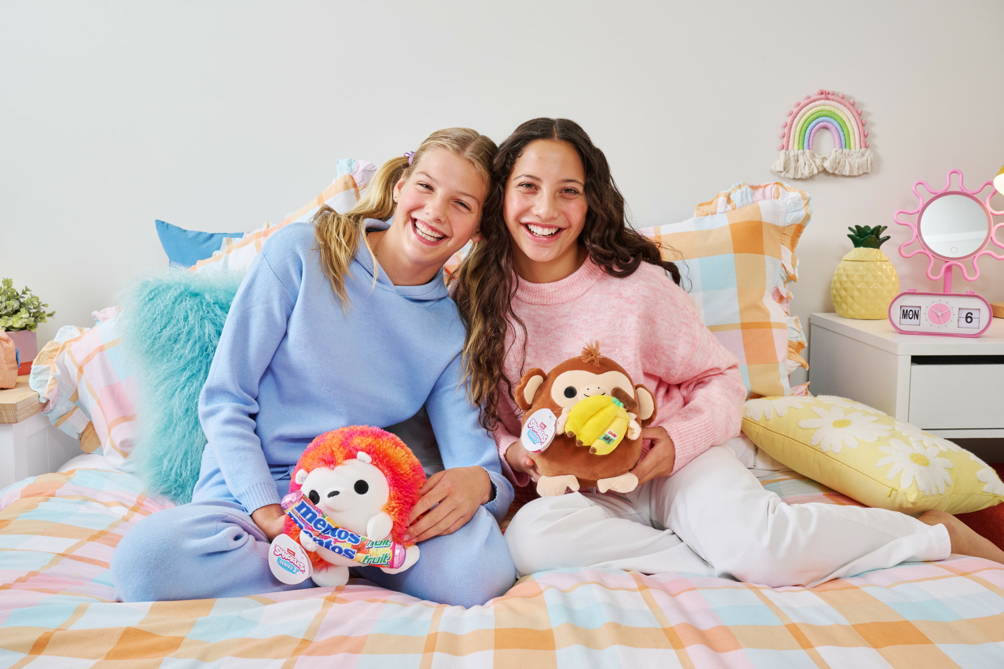 Zwei Mädchen lächeln, halten Plüschtiere. Eines mit Affe, das andere mit Igel. Bett mit bunten Laken.