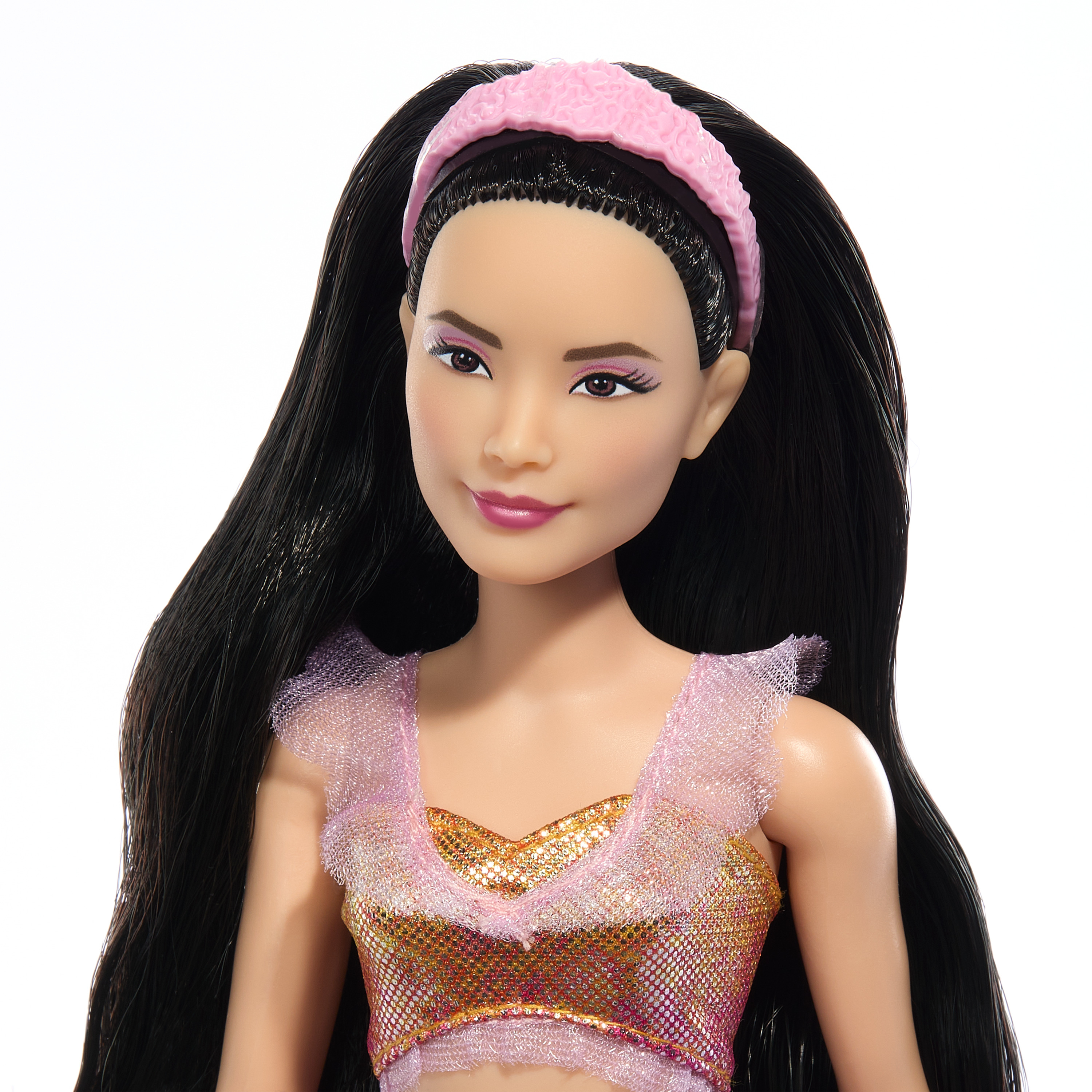 Nahaufnahme des Gesichts einer Puppe mit schwarzen Haaren, die ein rosa Stirnband und ein goldenes Oberteil trägt. Der Hintergrund ist weiß.