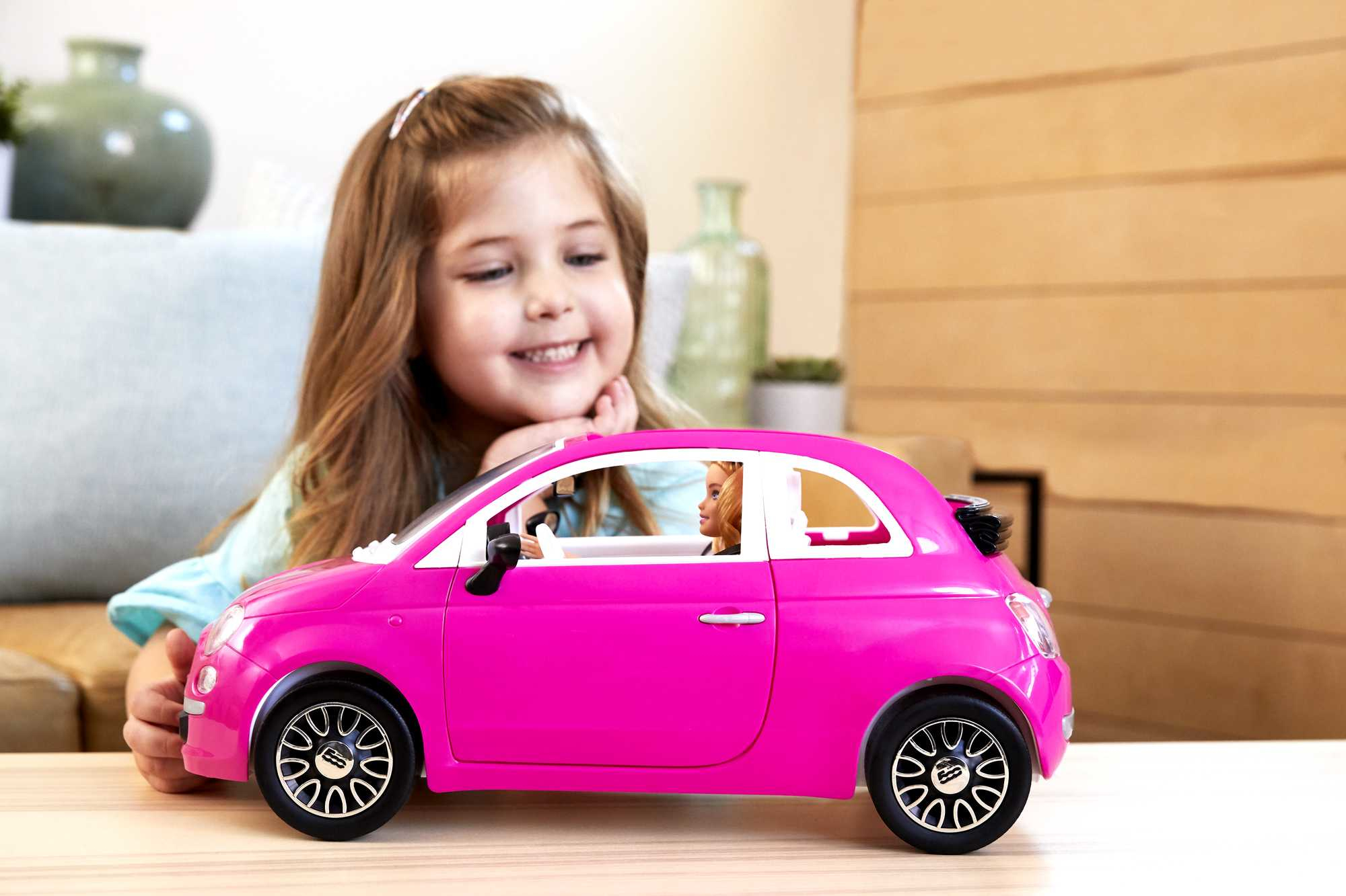 Ein Mädchen lächelt, während es ein pinkfarbenes Barbie-Auto mit einer Barbie-Puppe darin betrachtet.