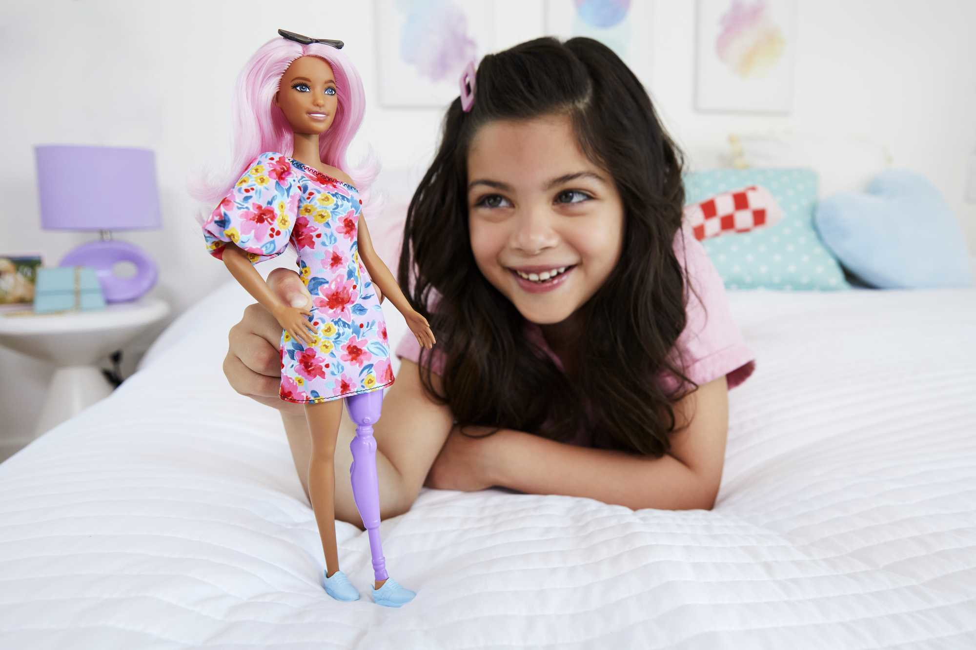 Ein Mädchen mit Barbie-Puppe. Die Puppe hat rosa Haare und ein Beinprothese. Weißer Hintergrund.