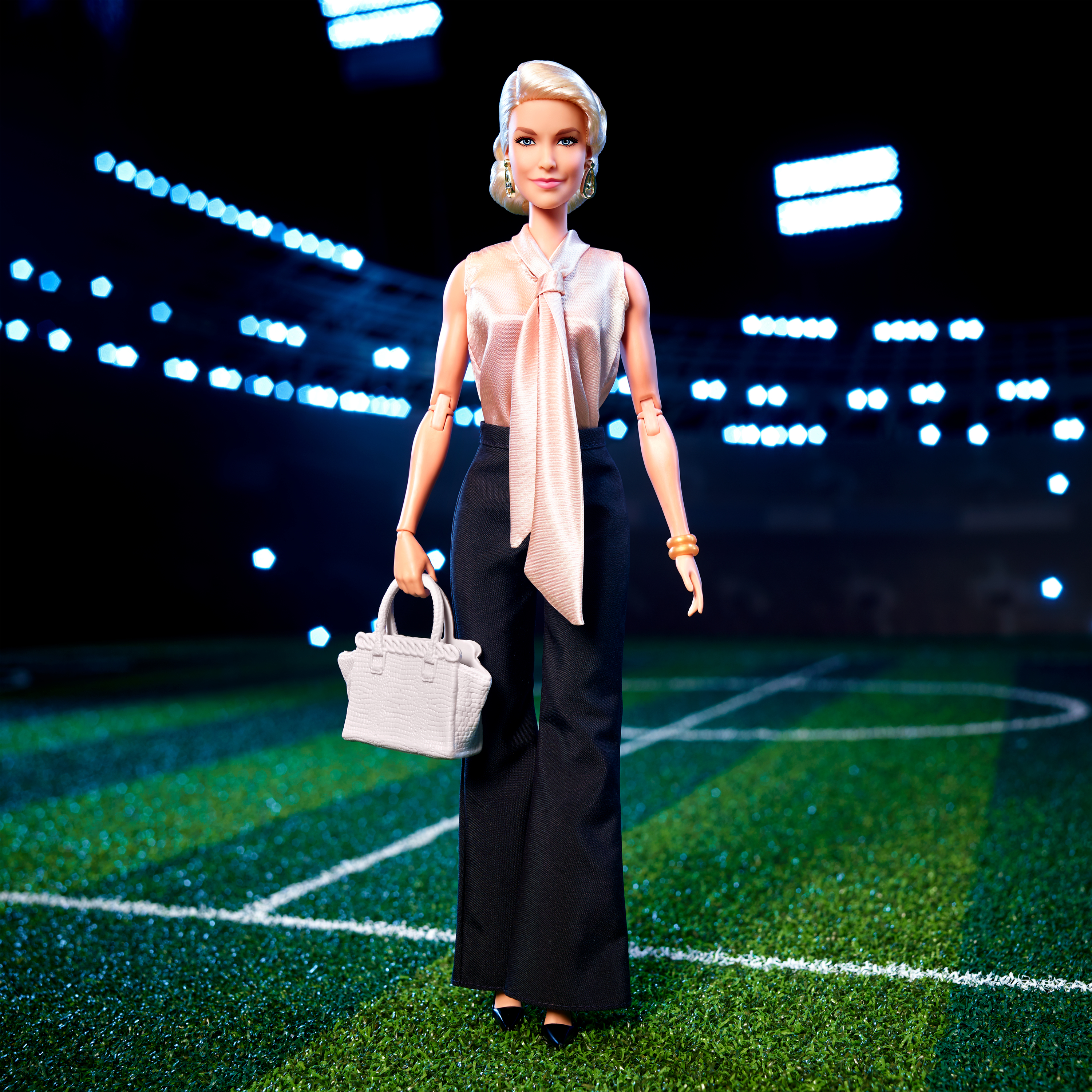 Blonde Barbie-Puppe in Business-Kleidung steht auf einem grünen Feld. Hintergrund: Stadionlichter.