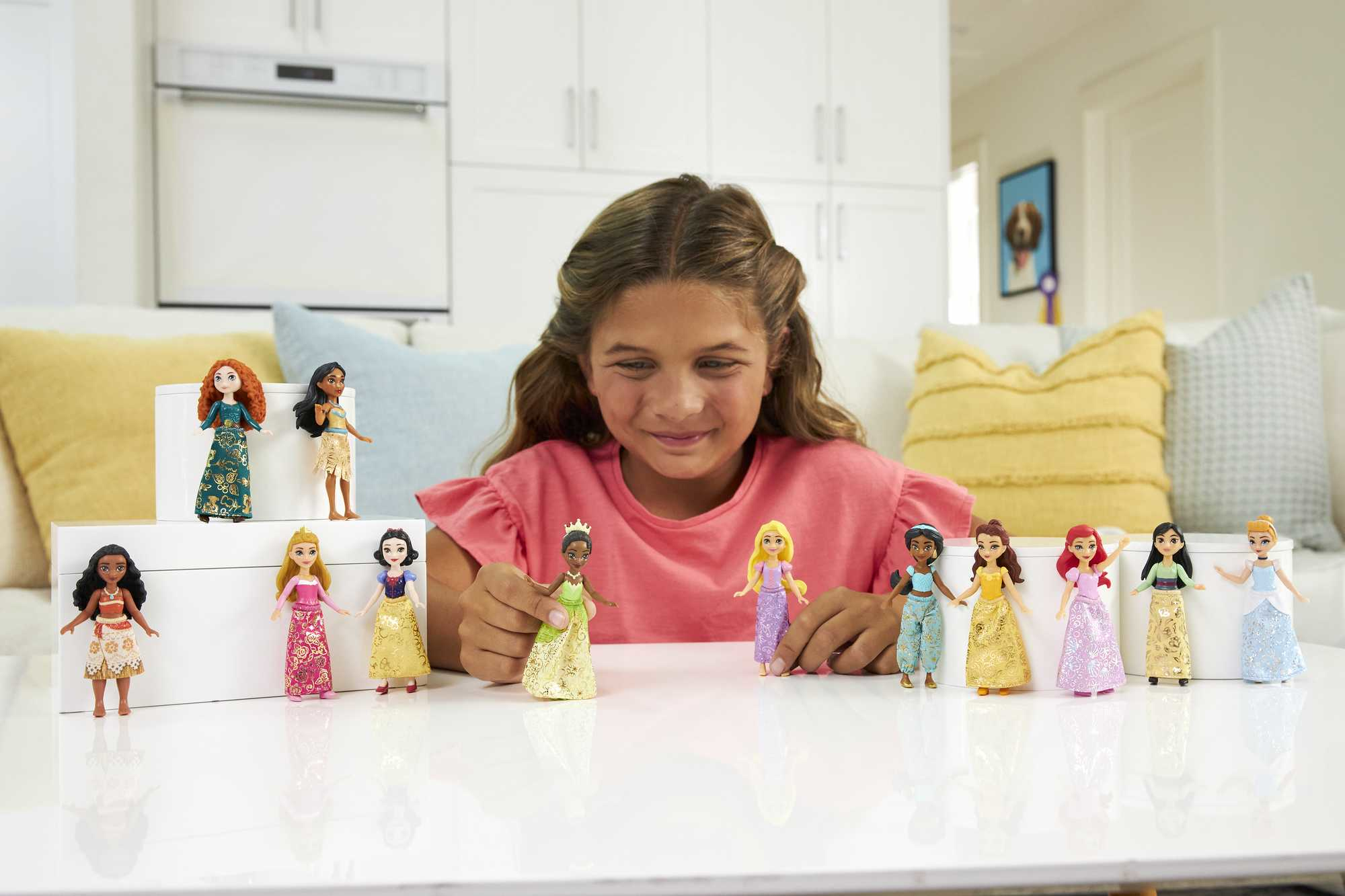 Ein lächelndes Mädchen, umgeben von verschiedenen Disney-Prinzessinnen-Puppen auf einem Tisch.