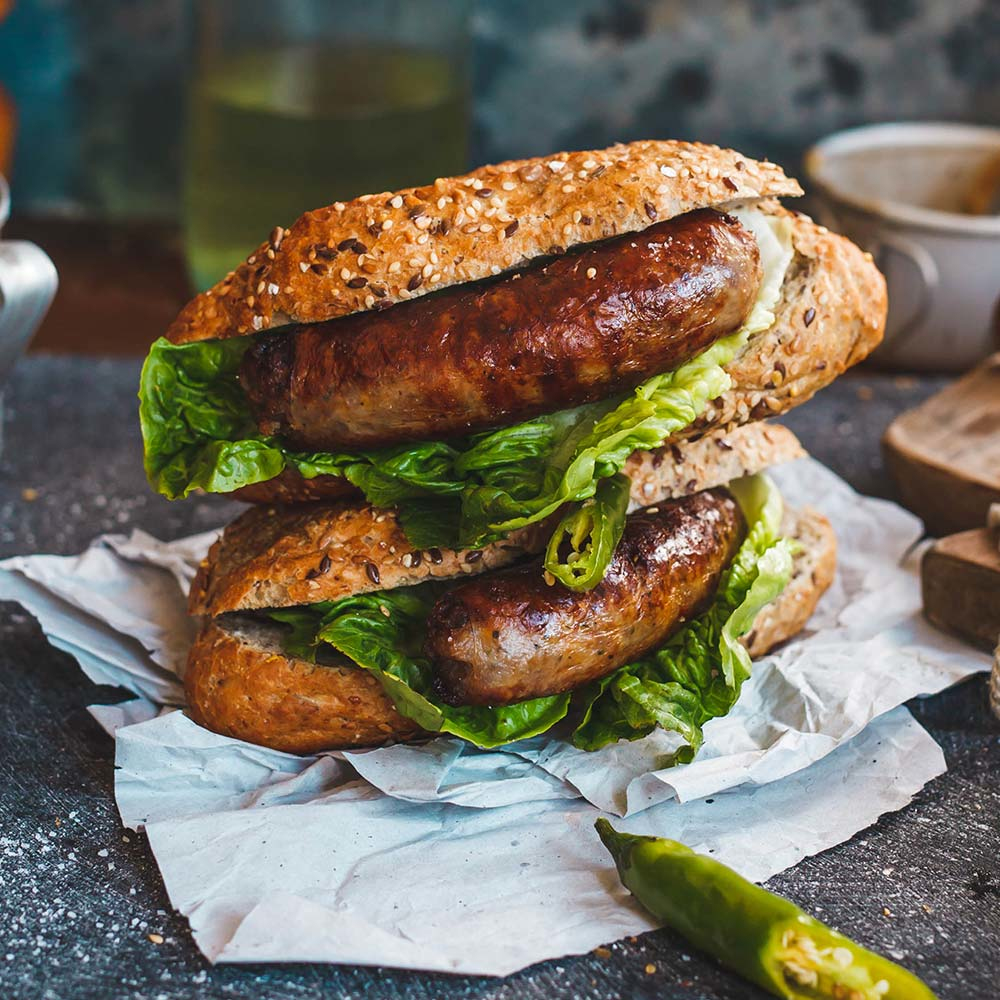 Due panini con salsiccia con pane ai semi di sesamo. I panini hanno lattuga e le salsicce sono dorate.